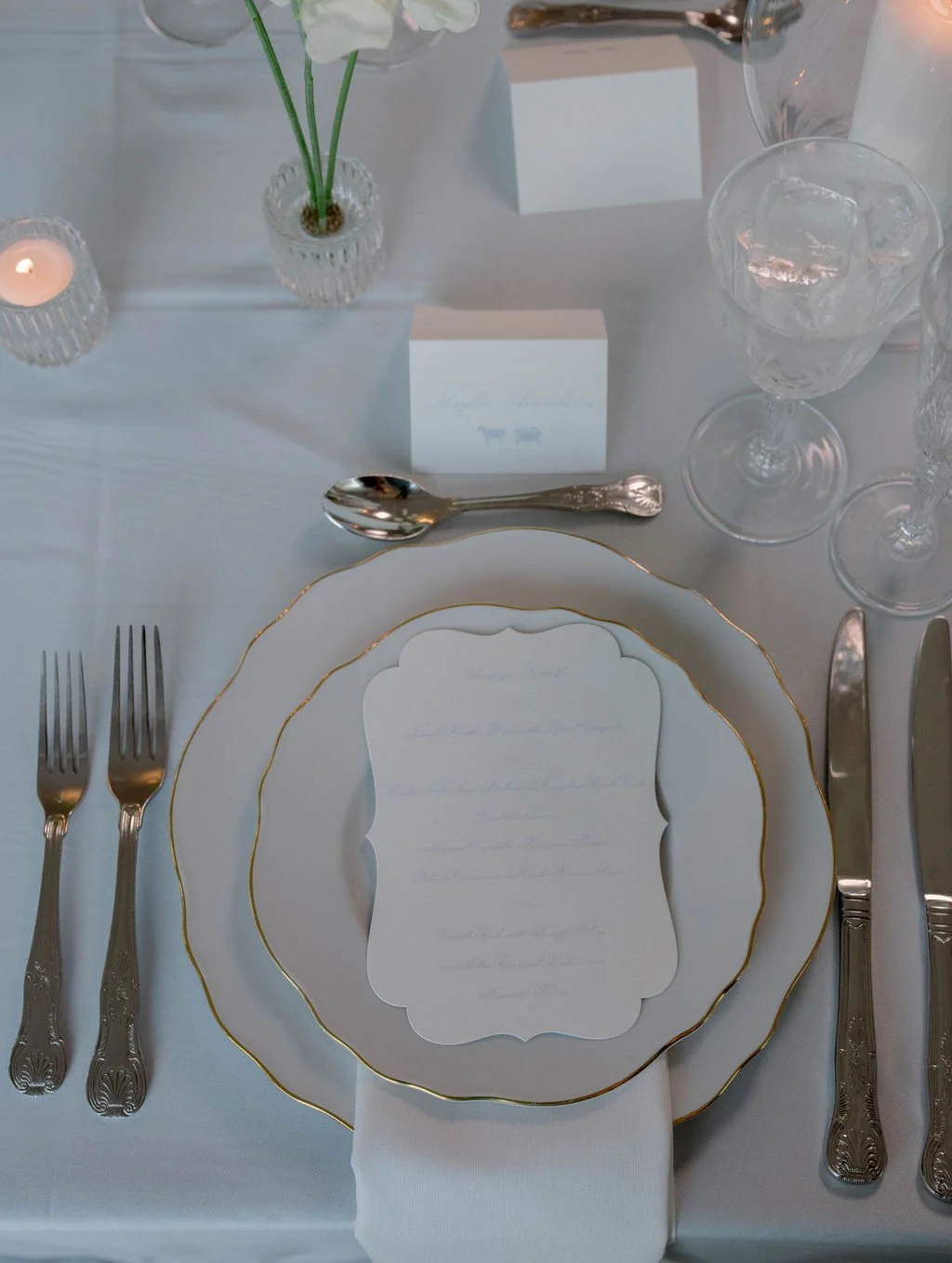 Elegant place setting with white plates with gold trim, a menu card, silverware, glassware with ice, a small flower vase with white flowers, and a lit candle on a white tablecloth.