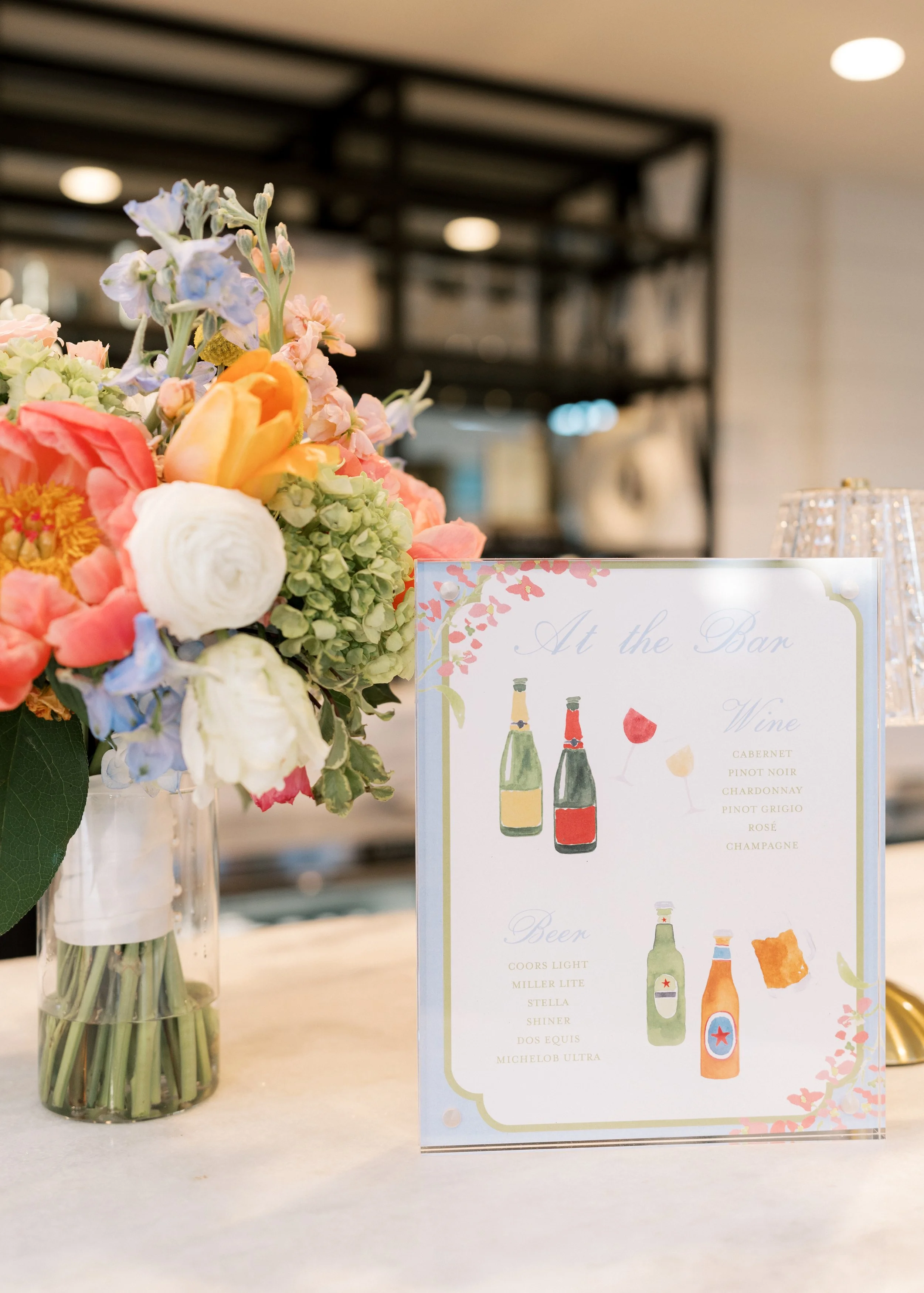 A floral arrangement in a glass vase positioned on a white table next to a drink menu with watercolor-style illustrations of wine and beer bottles and glasses.