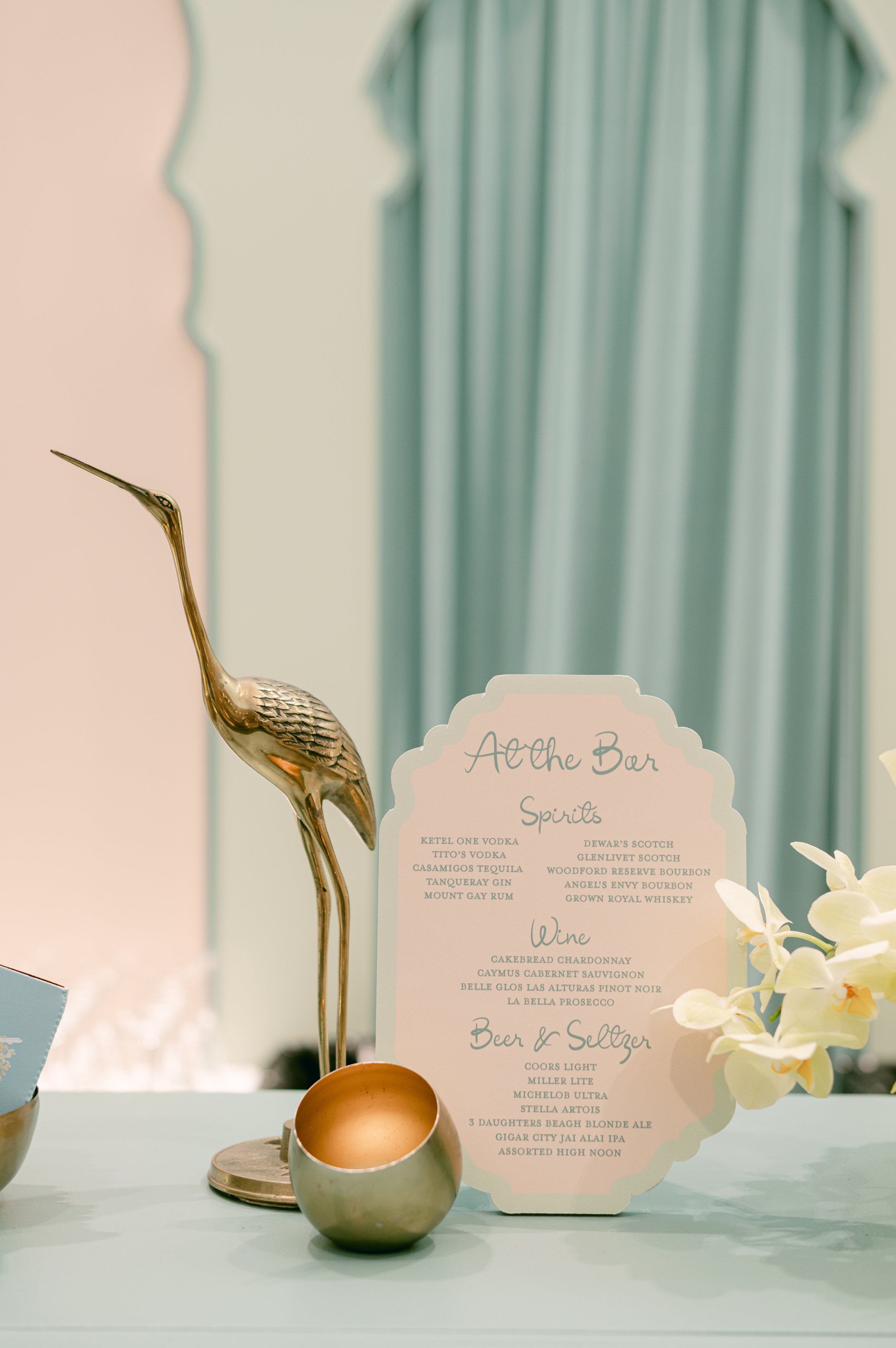 A pastel-colored bar setup with a decorative golden heron statue, a gold and silver container, a flower arrangement, and a menu titled 'Aloha Bar' listing spirits, wines, beers, and seltzers.