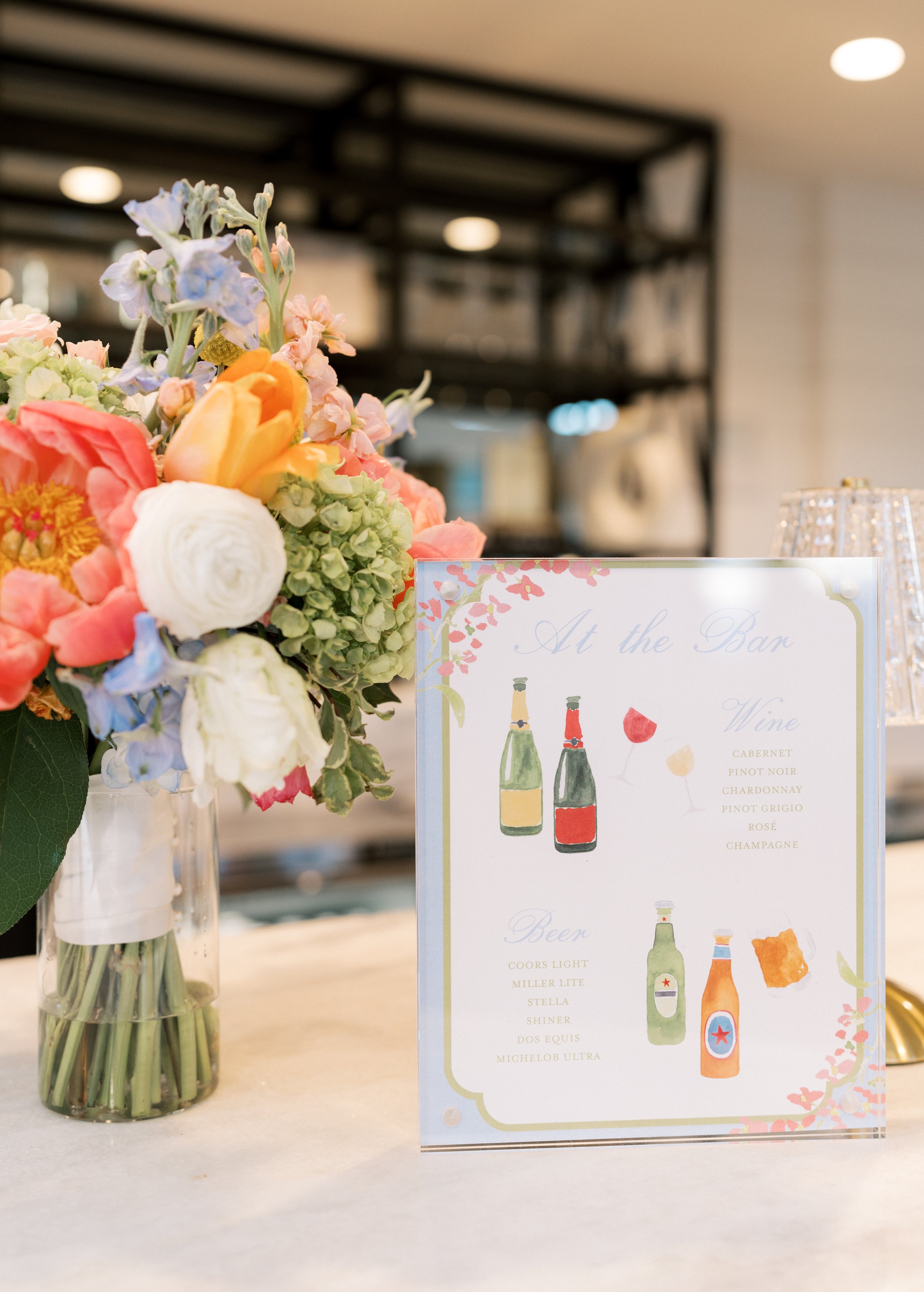A colorful flower arrangement featuring pink, white, and blue flowers on a table, next to a wine and beer menu with illustrated bottles and glasses.