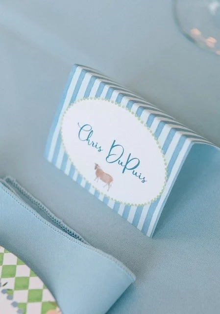 Blue striped card with the name 'Chris Dupuis' and a small illustration of a cow, placed on a light blue surface with a zipper and part of a patterned fabric visible.