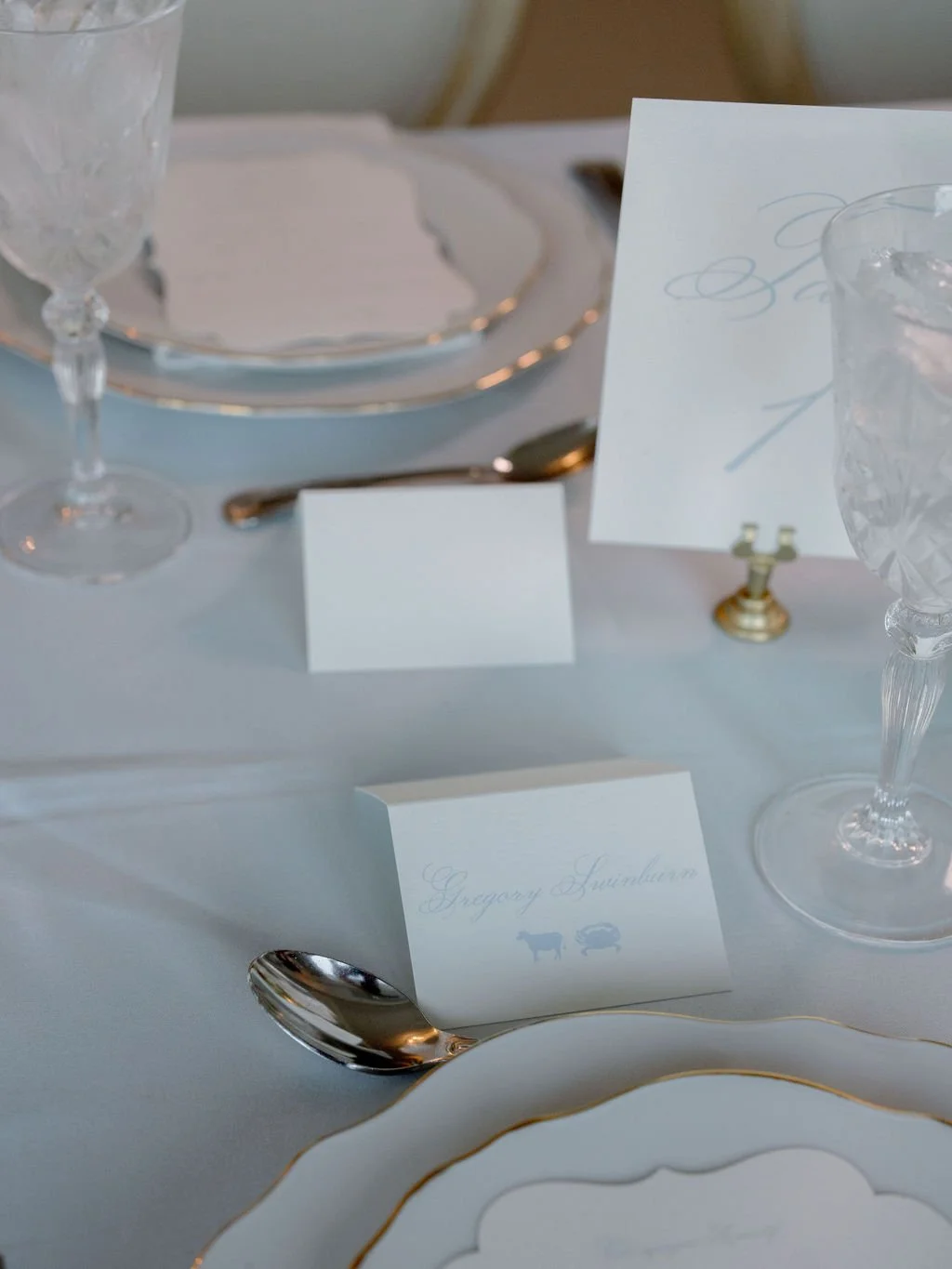 Elegant table setting with white plates, crystal glasses, a spoon, and place cards, including one that reads 'Gregory Lyonbaum' with animal icons, on a white tablecloth with gold accents.