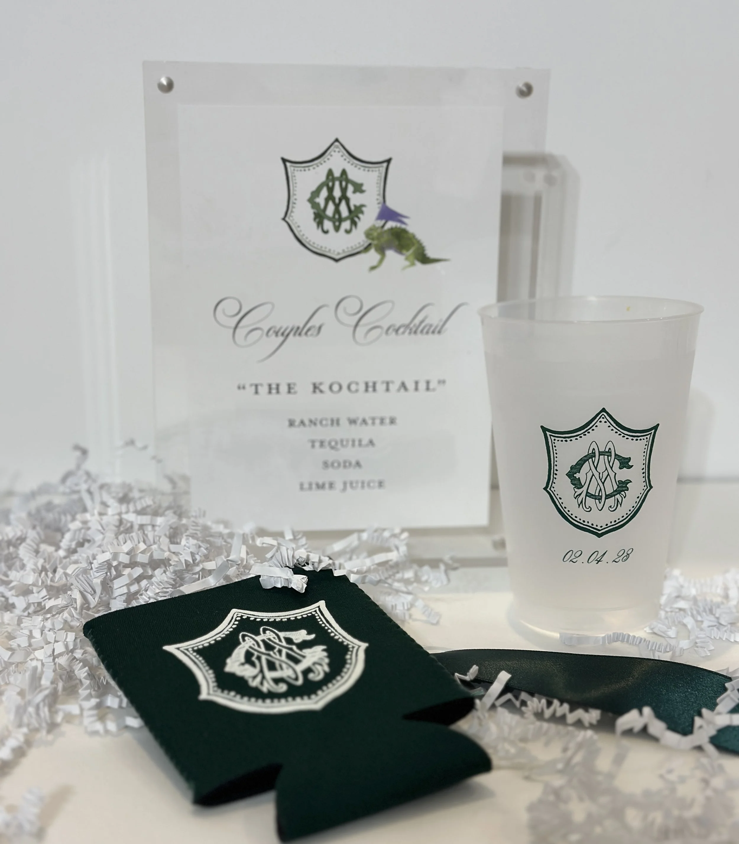 A clear plastic cup with a dragon and crest logo, a black cloth with a similar crest, a printed menu with crest and a dragon, all arranged among white shredded paper.