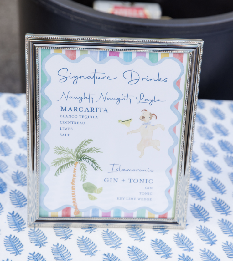 A colorful framed sign on a table with a blue leaf pattern tablecloth, listing signature drinks including a Margarita with ingredients and an Islamoranie Gin and Tonic.