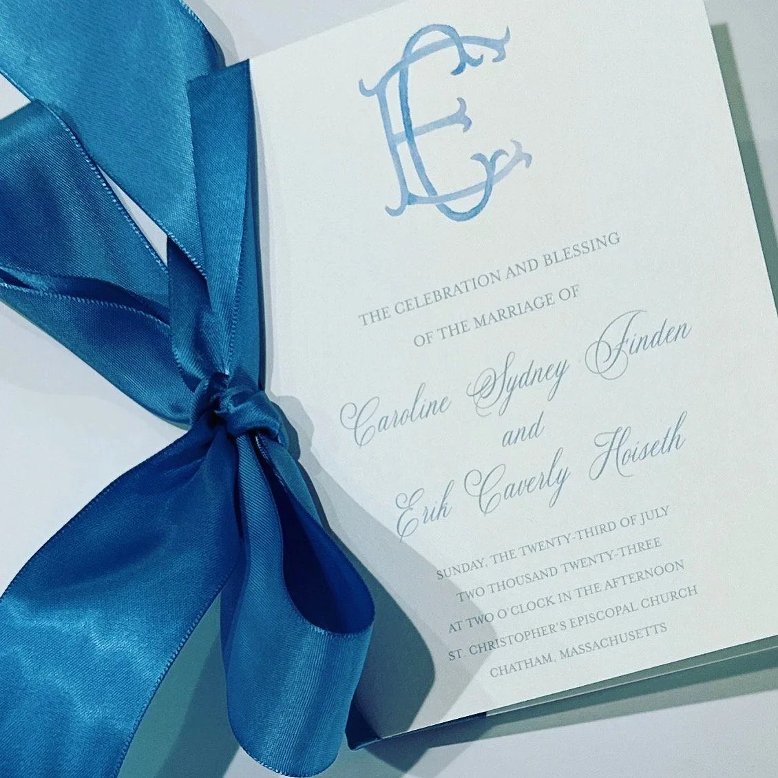 Wedding invitation with a blue ribbon, featuring a monogram at the top and details of the marriage of Caroline Sydney Junden and Erik Caverly Houseth at St. Christopher's Episcopal Church in Chatham, Massachusetts.