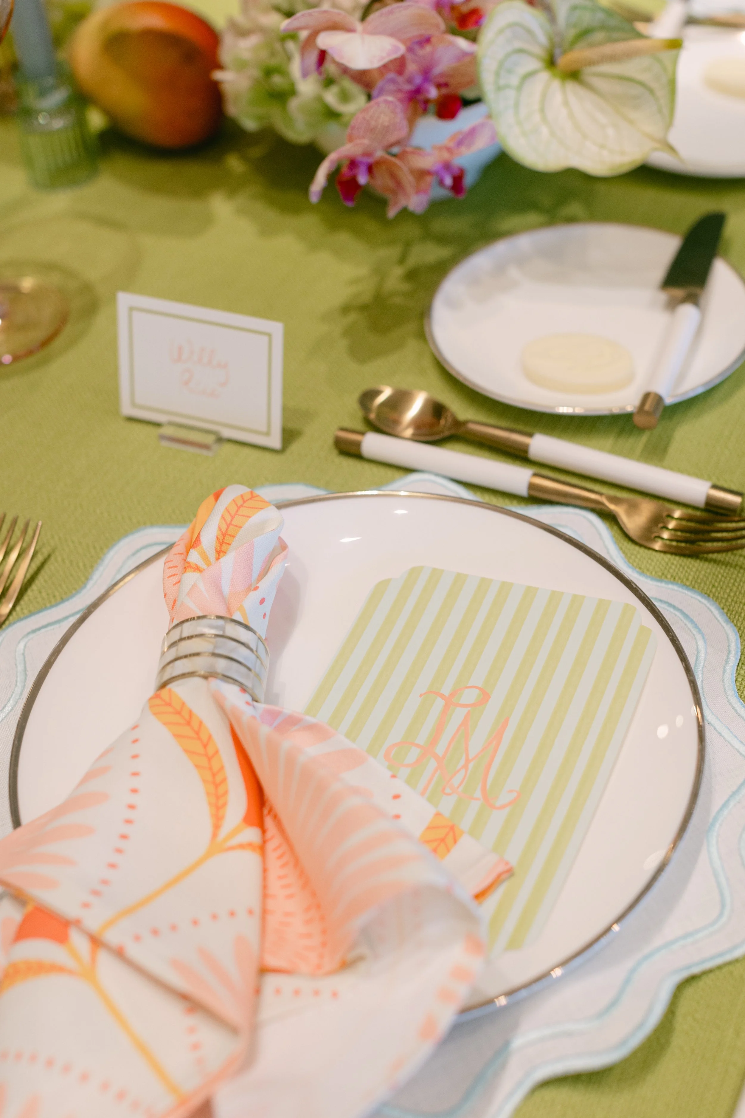A table setting with a white plate, a pink and orange napkin held by a silver napkin ring, a green and white striped card with pink monogram initials, gold cutlery, a small white plate with a butter knife, a green tablecloth, and a floral centerpiece