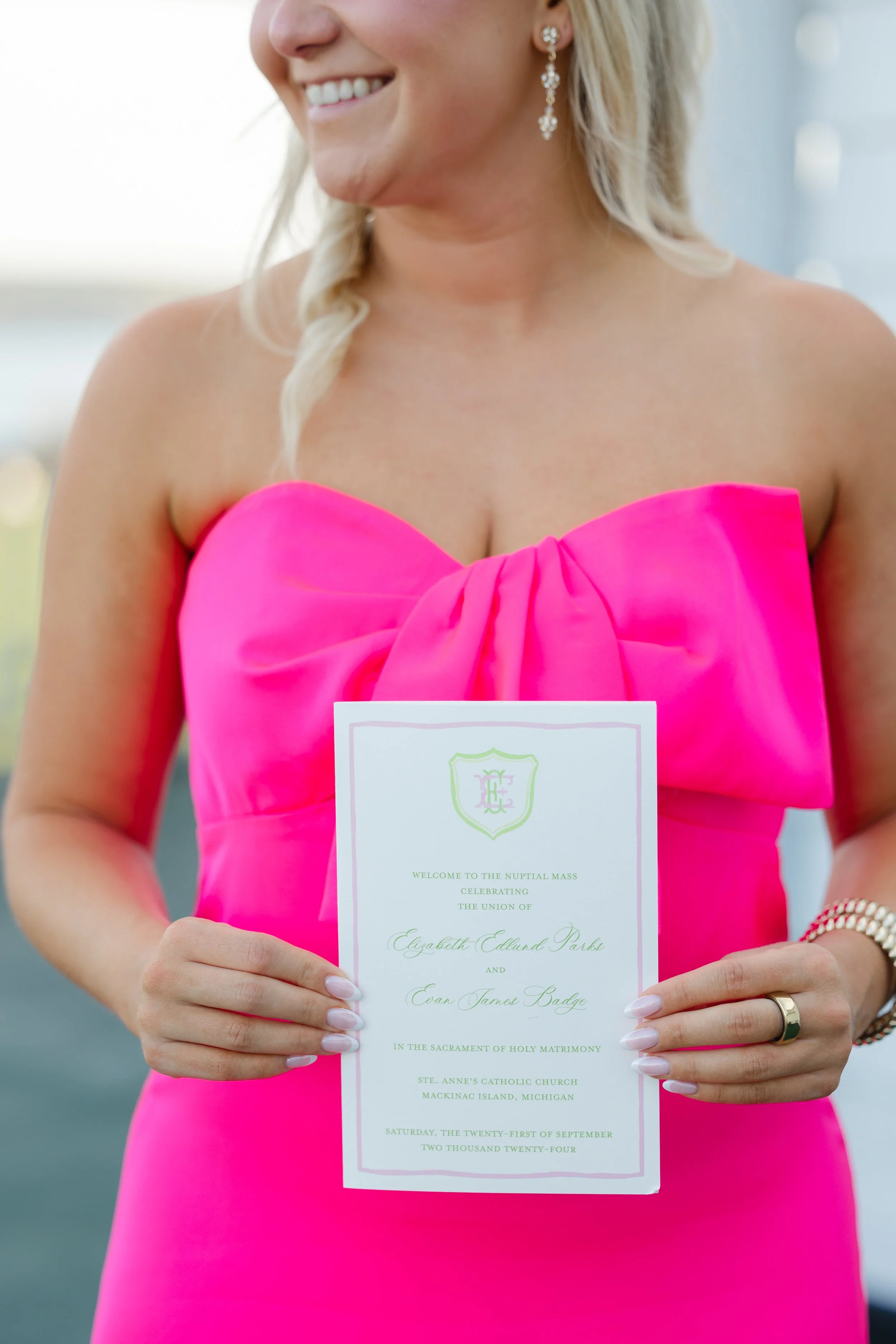 Woman in a bright pink strapless dress holding a wedding program, smiling and standing outdoors.