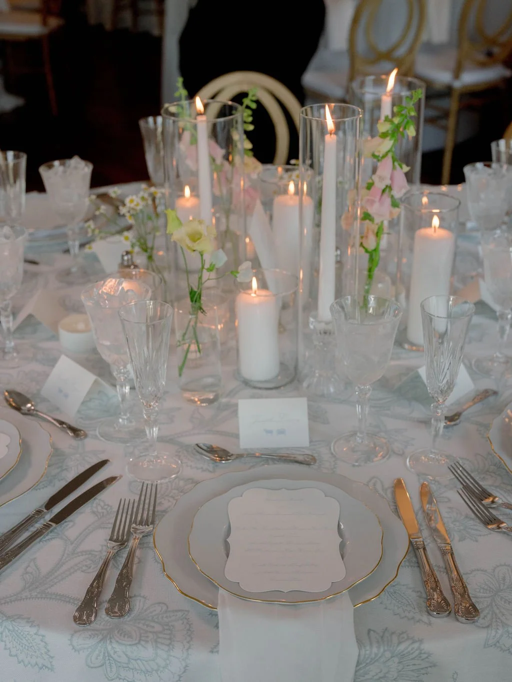 Elegant round table set for a formal event with white and gold-rimmed dishes, silverware, clear glassware, and a centerpiece of tall white candles and pink and white flowers at a wedding or banquet.