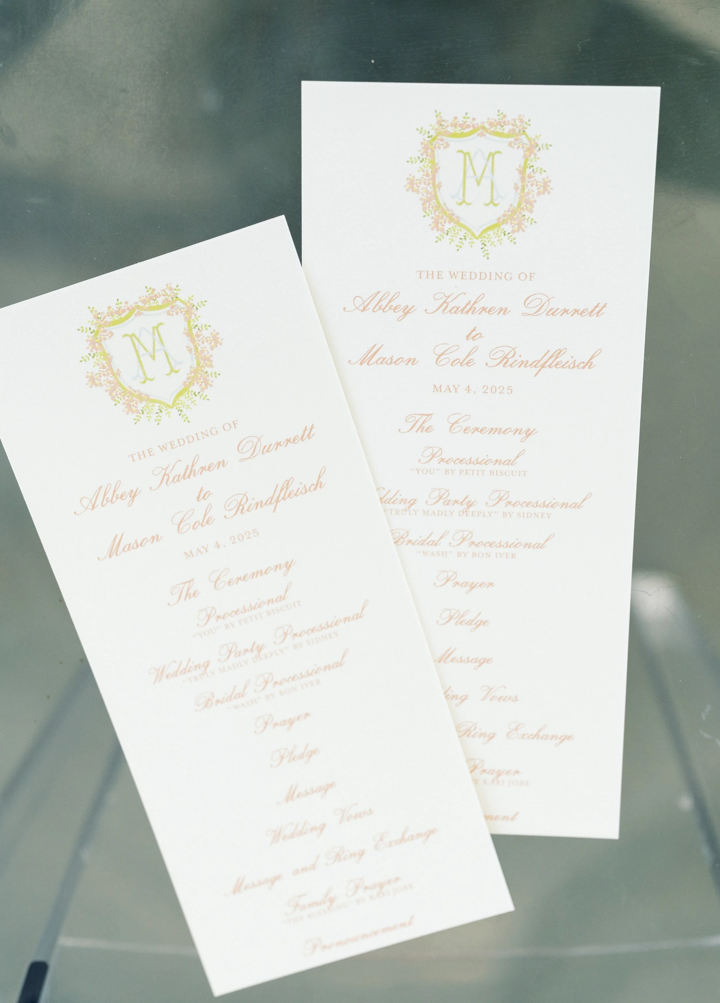 Two white wedding programs with pastel floral crest and pink cursive text, listing ceremony and reception events, on a glass table.