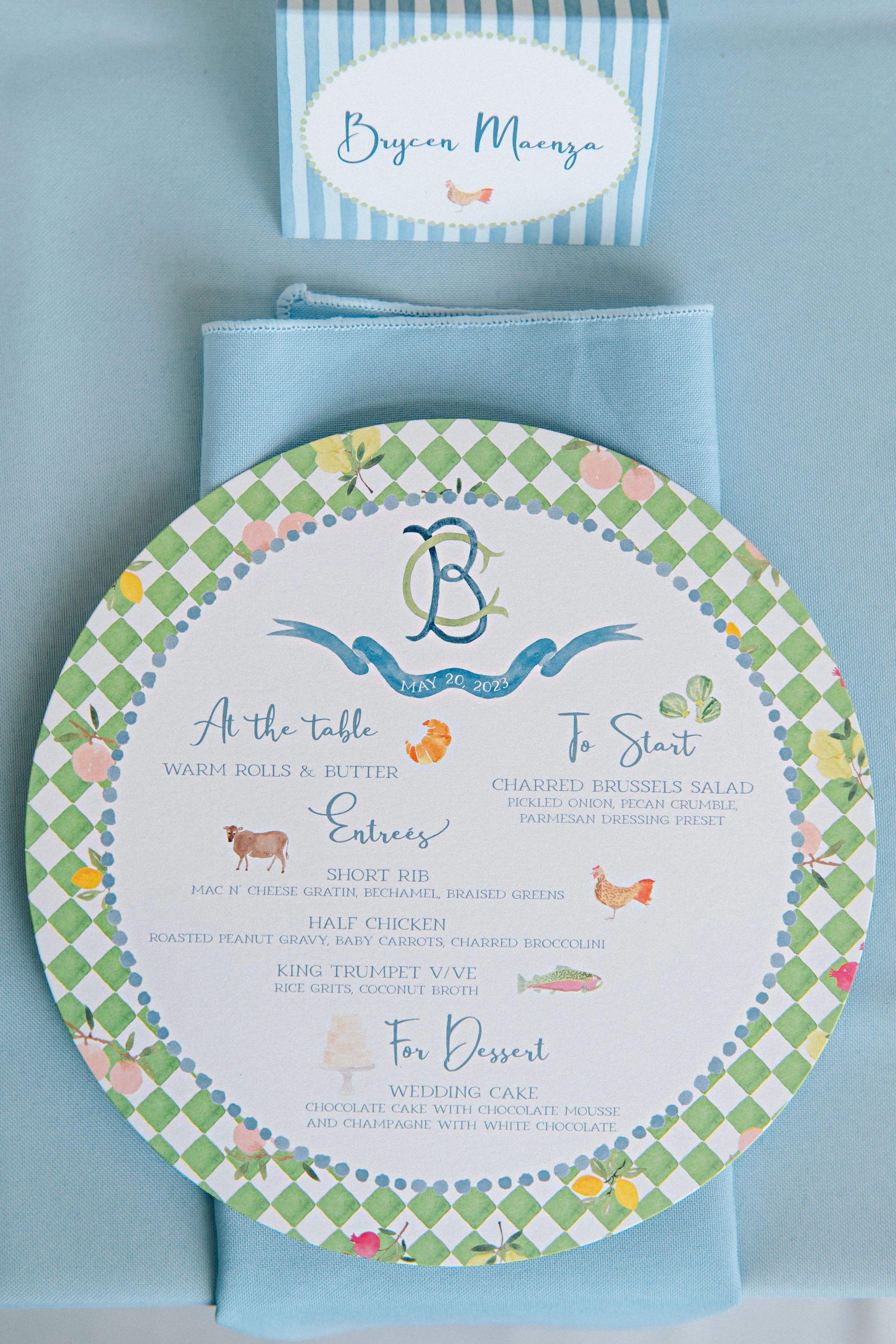 Wedding reception menu with a lemon-themed border, featuring a monogram and date, listing a meal of warm rolls and butter, charred Brussels salad, short rib, half chicken, and king trumpet veal, with wedding cake for dessert, set on a light blue tabl
