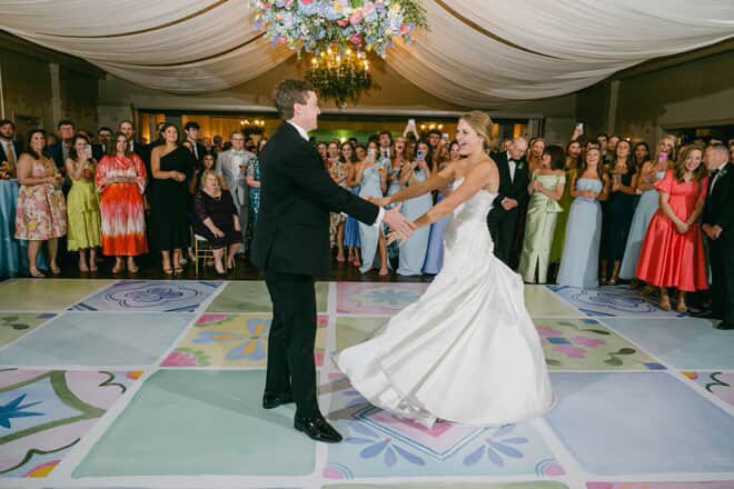 A bride and groom dancing at their wedding reception with guests watching in a decorated indoor venue.