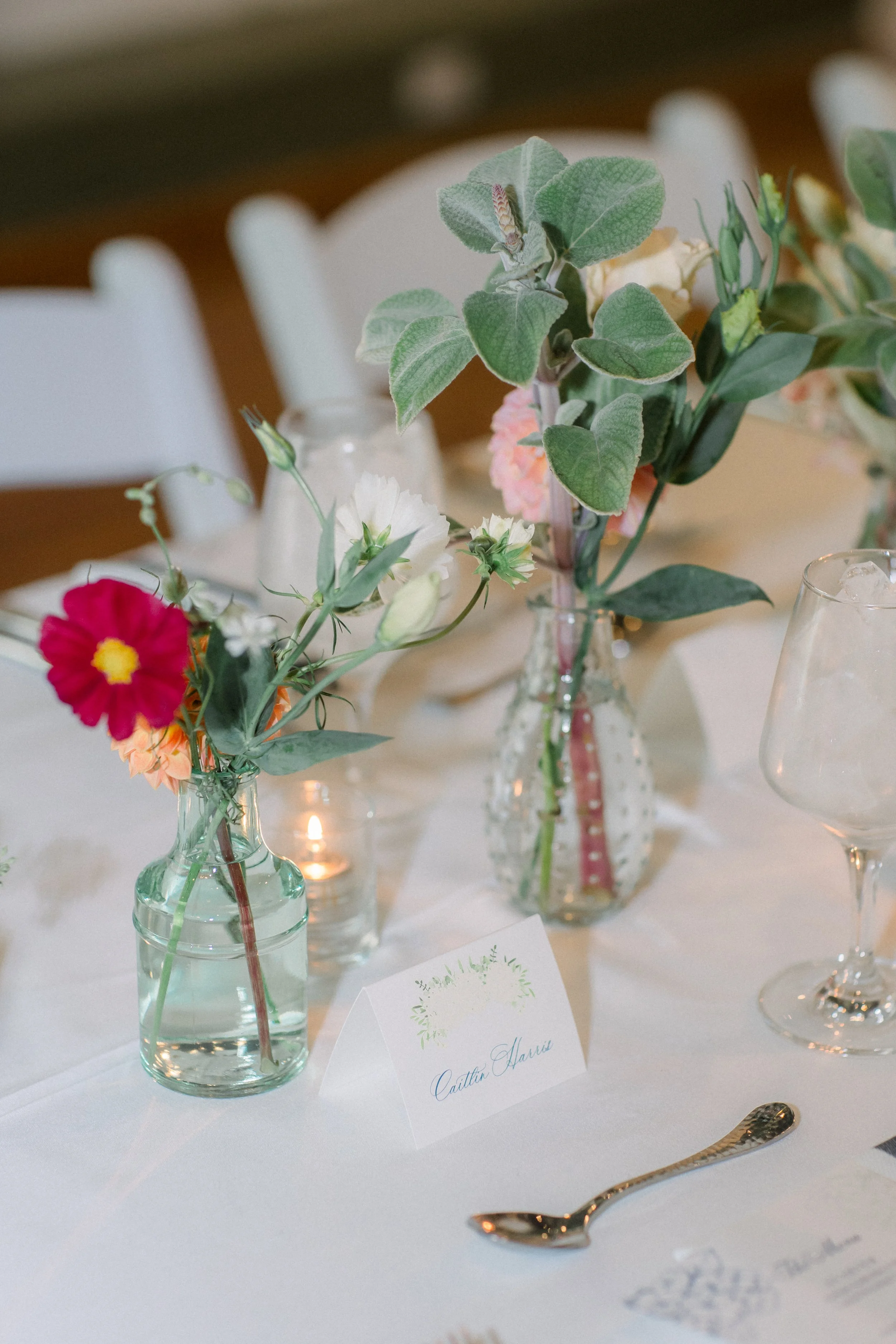 Table centerpiece with assorted flowers in small glass vases and a place card with the name Caitlin Harris, along with a candle and a gold spoon, set on a white tablecloth at a formal event.