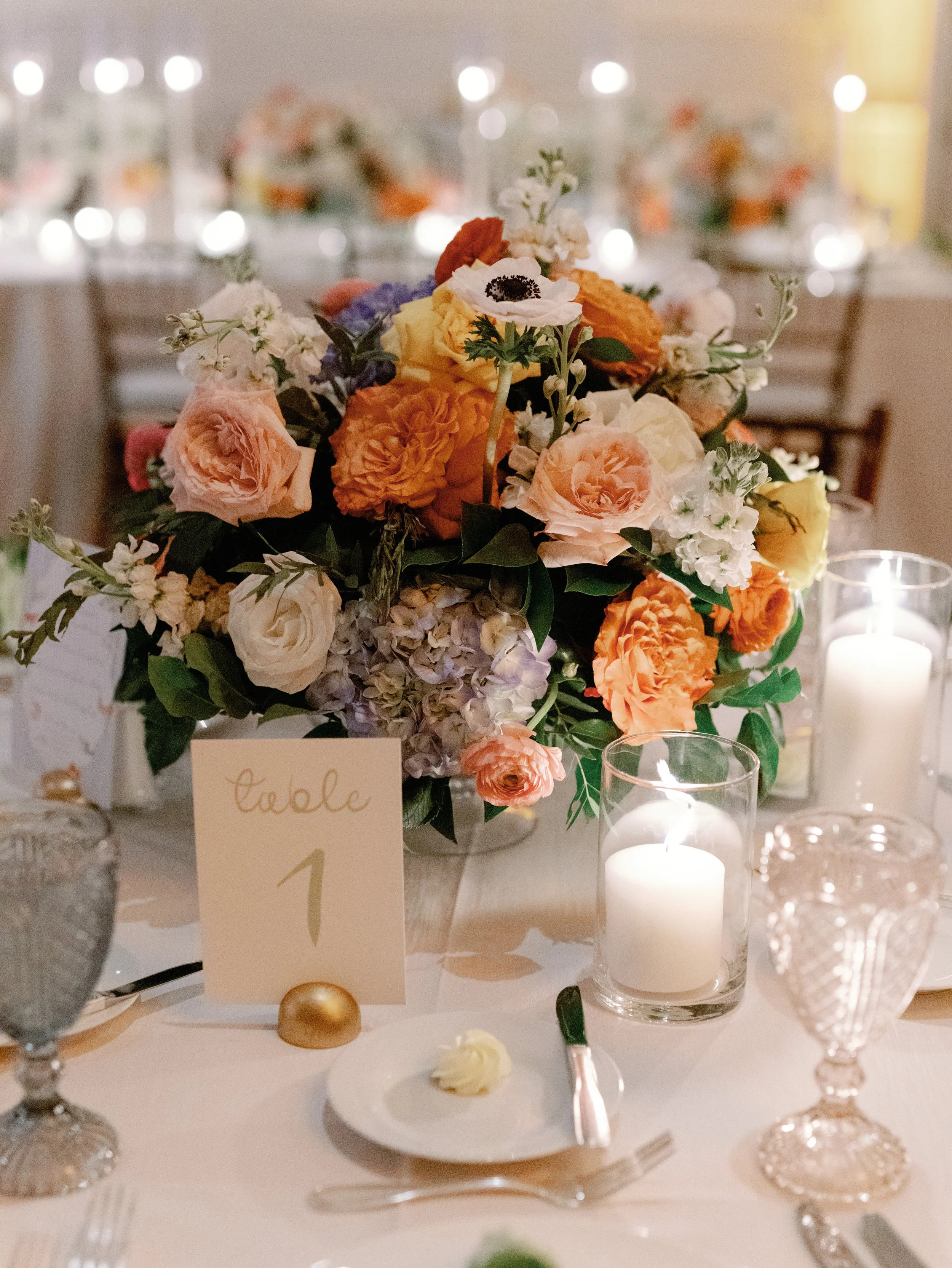 Elegant table centerpiece with a colorful flower arrangement, candles, and a table number card at a wedding reception.