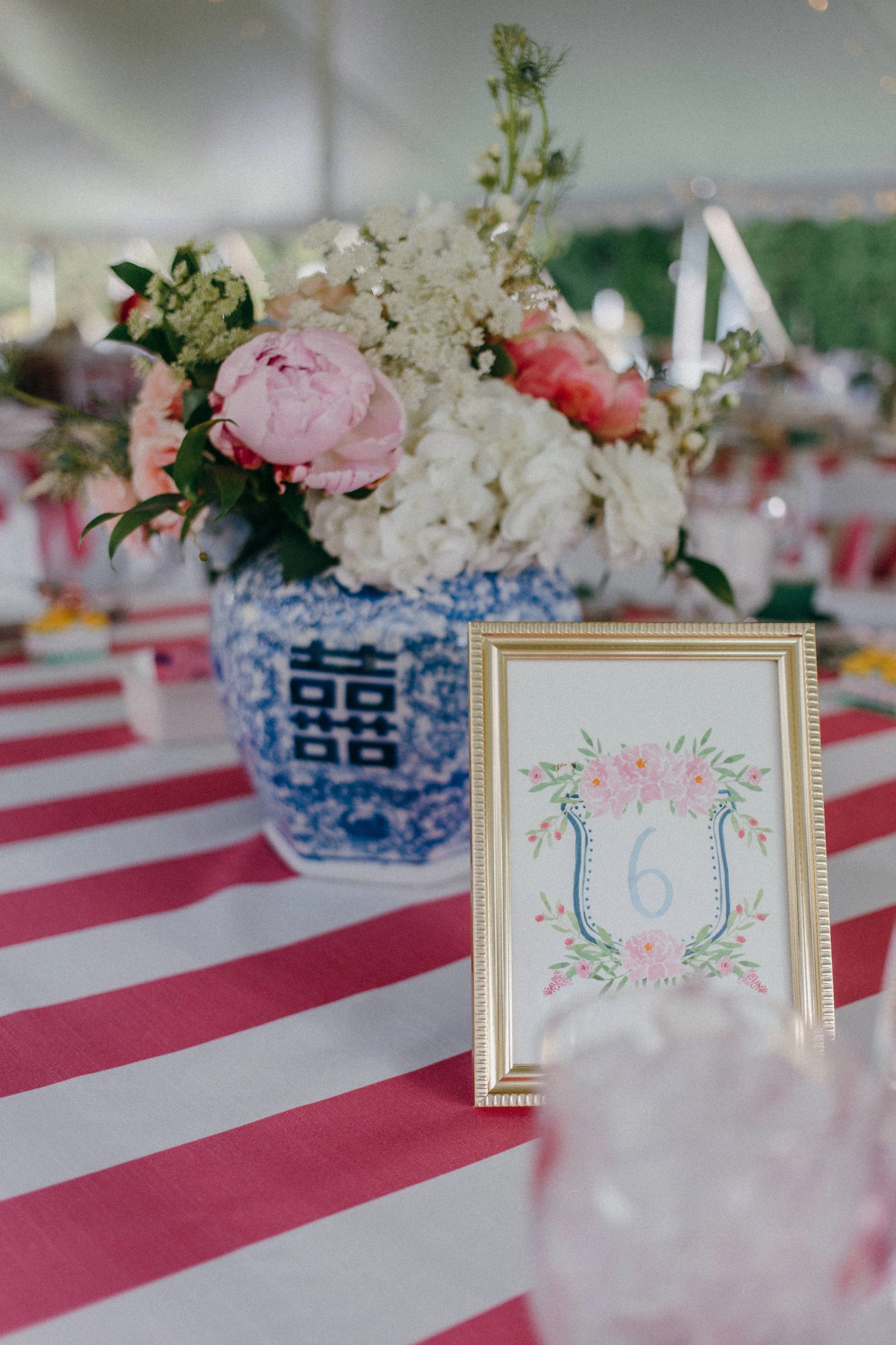 A table centerpiece with a bouquet of pink and white flowers in a blue and white patterned vase, with a framed table number six decorated with pink flowers and green leaves, on a table with a red and white striped tablecloth at an outdoor event.