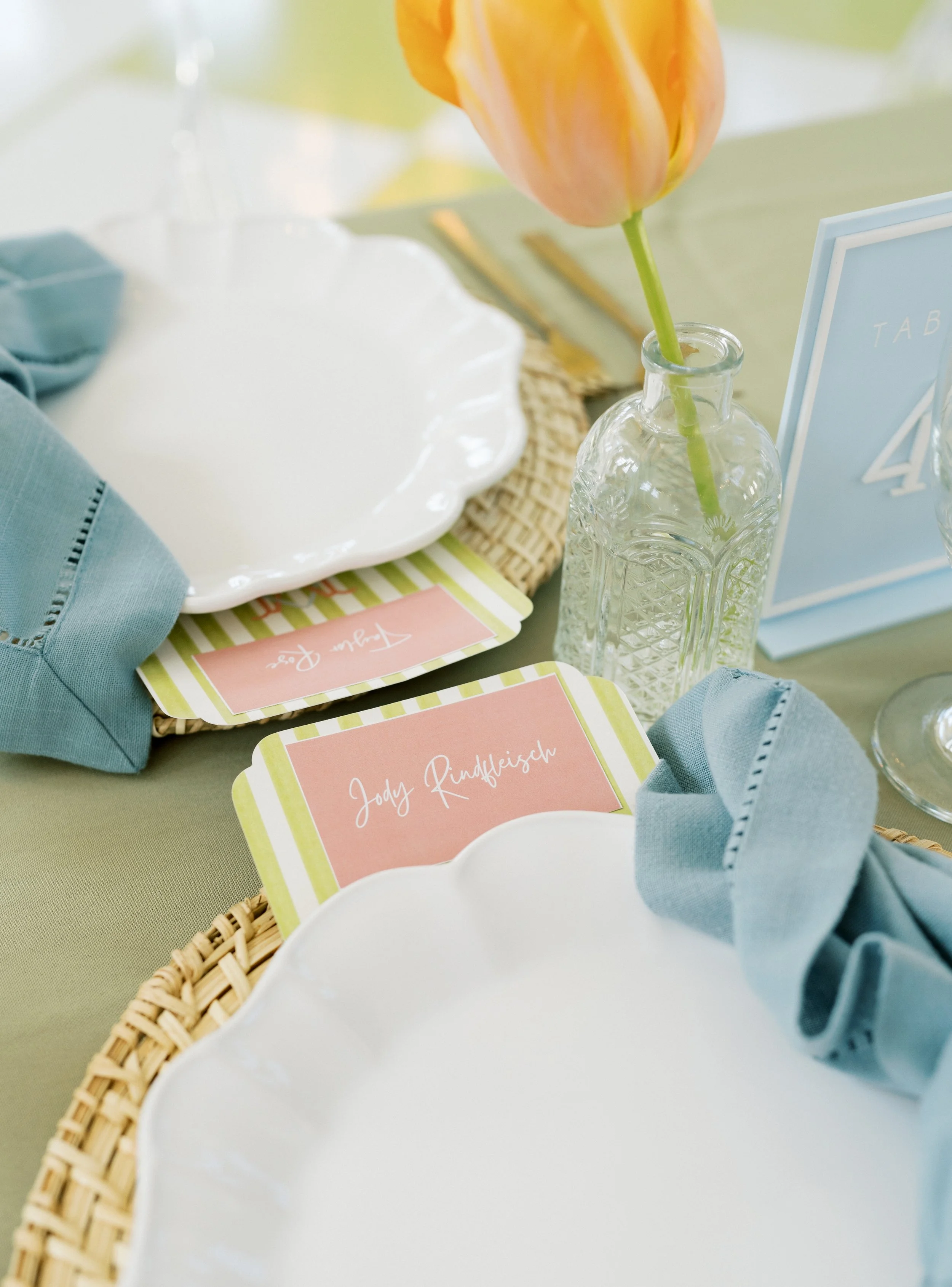 A table setting with a clear glass bottle holding a orange and yellow tulip, white plates, folded blue napkins, table number card, and place card with the name Jody Rindfleisch on it, all arranged on a green tablecloth.