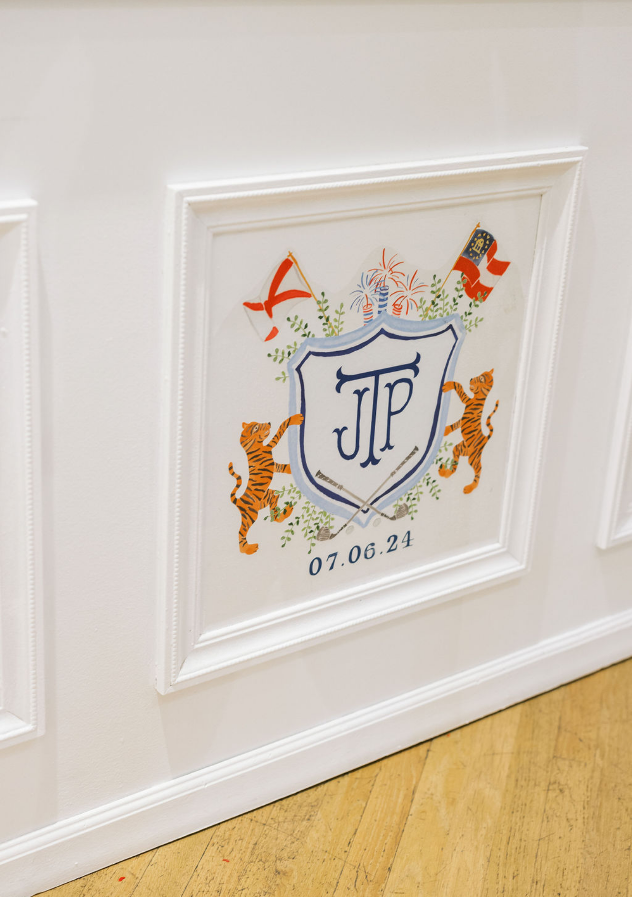 Decorative crest with initials JP, the date 07.06.24, two tigers, flags, colorful fireworks, and greenery on a white wall.