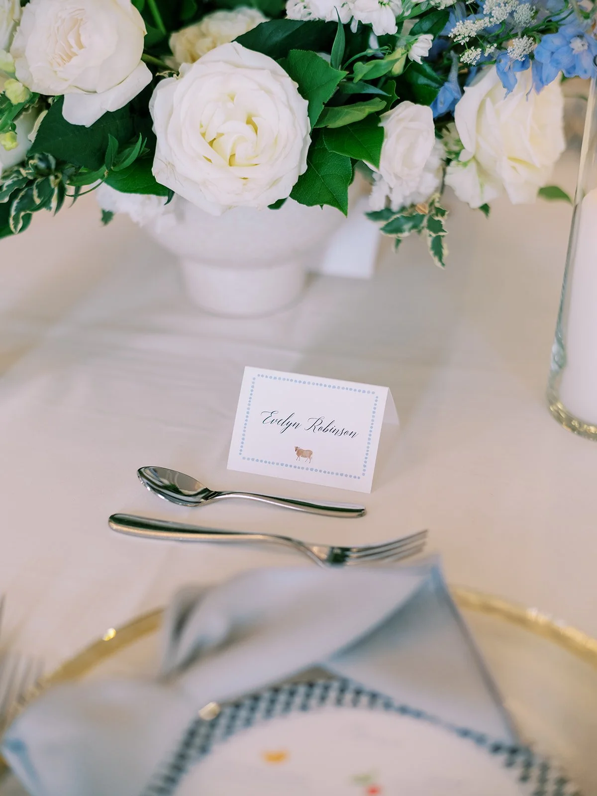 A white flower arrangement with roses and greenery on a table. A place card with the name Evelyn Robinson and a small cow illustration. Silver spoon and fork, folded napkin, and a candle in a glass holder on the table.