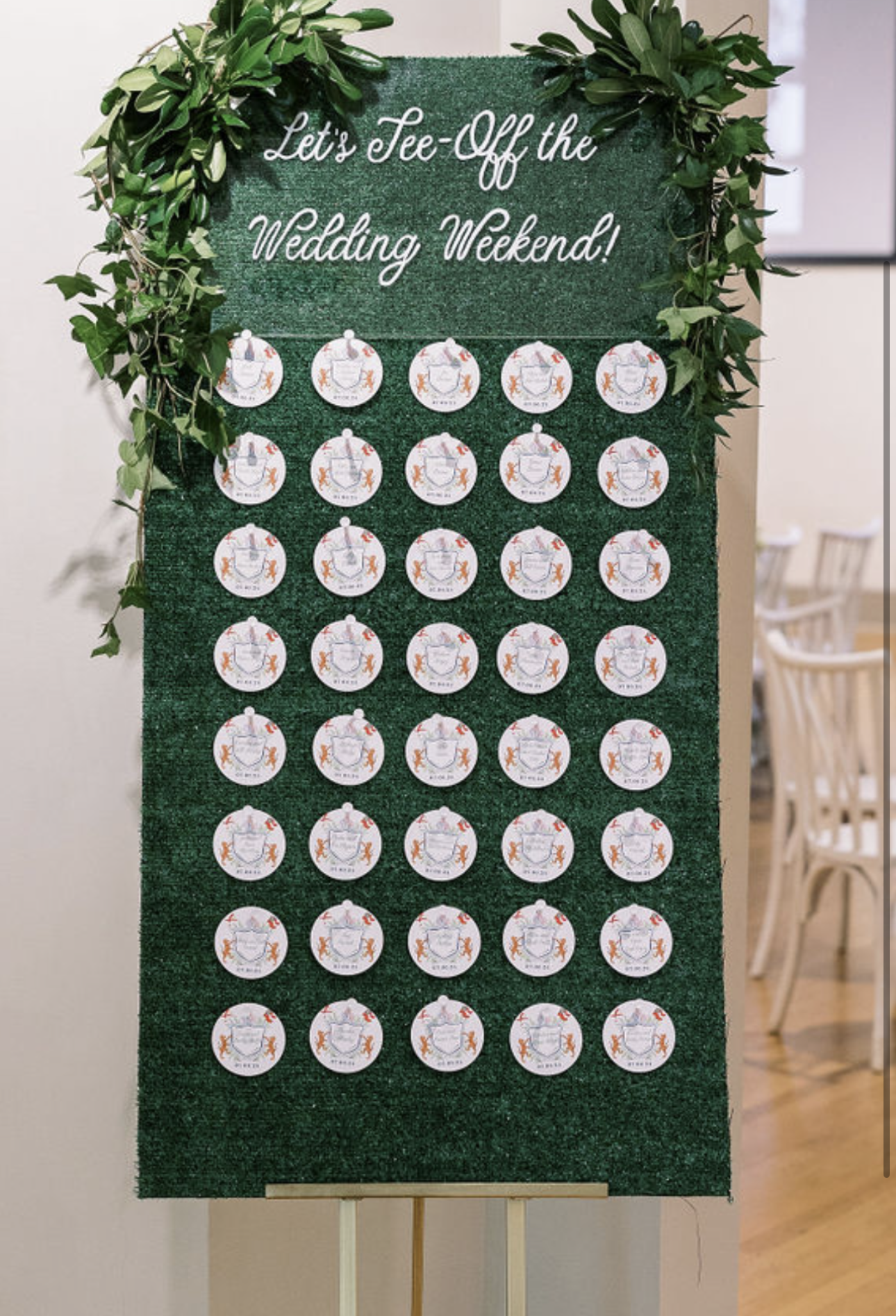 A green felt board decorated with green leaves at the top, with the white text "Let's Tee-Off the Wedding Weekend!" written in cursive. Several golf-themed name tags are arranged in rows below the text, each featuring a golf ball design and a golfer'