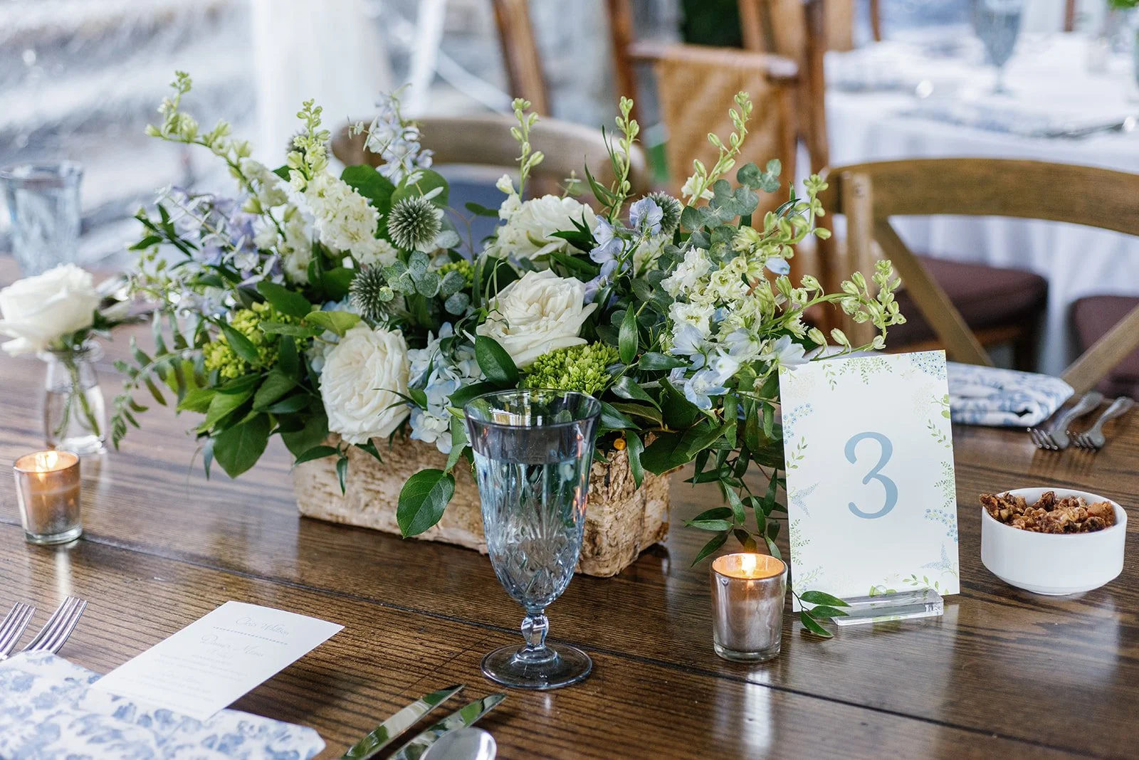 Centered floral arrangement of white roses, green foliage, and light blue flowers on a wooden table, with table number 3, candles, water glass, and set tableware.