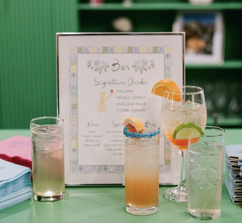 Four glasses of signature drinks with garnishes on a green table in front of a menu sign.