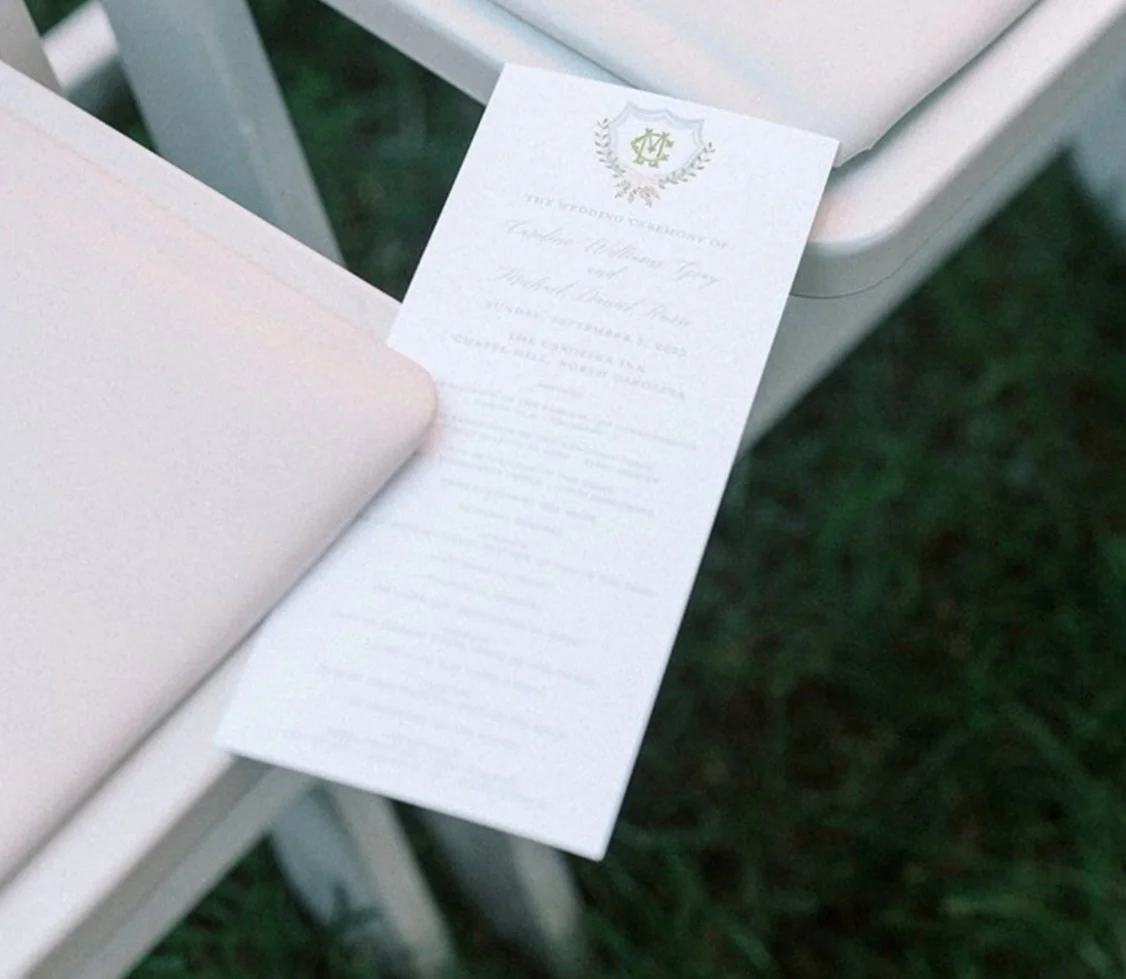 A wedding invitation printed on white paper with a gold emblem, placed on a white bench outdoors with grass in the background.