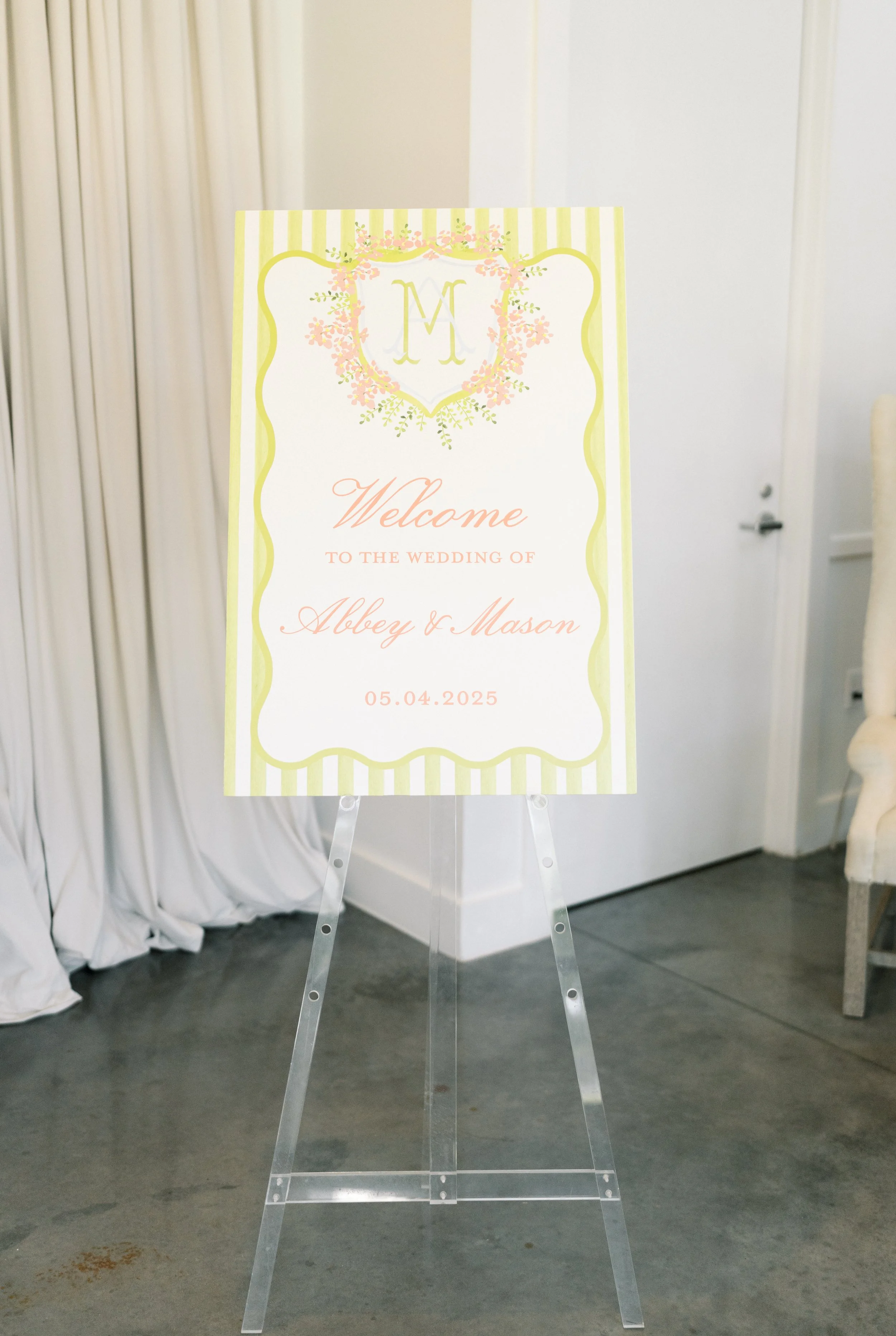 Wedding welcome sign with a yellow and pink floral design, featuring the initials 'M' at the top, and text welcoming guests to the wedding of Abbey and Mason on May 4, 2025, on a decorative stand.