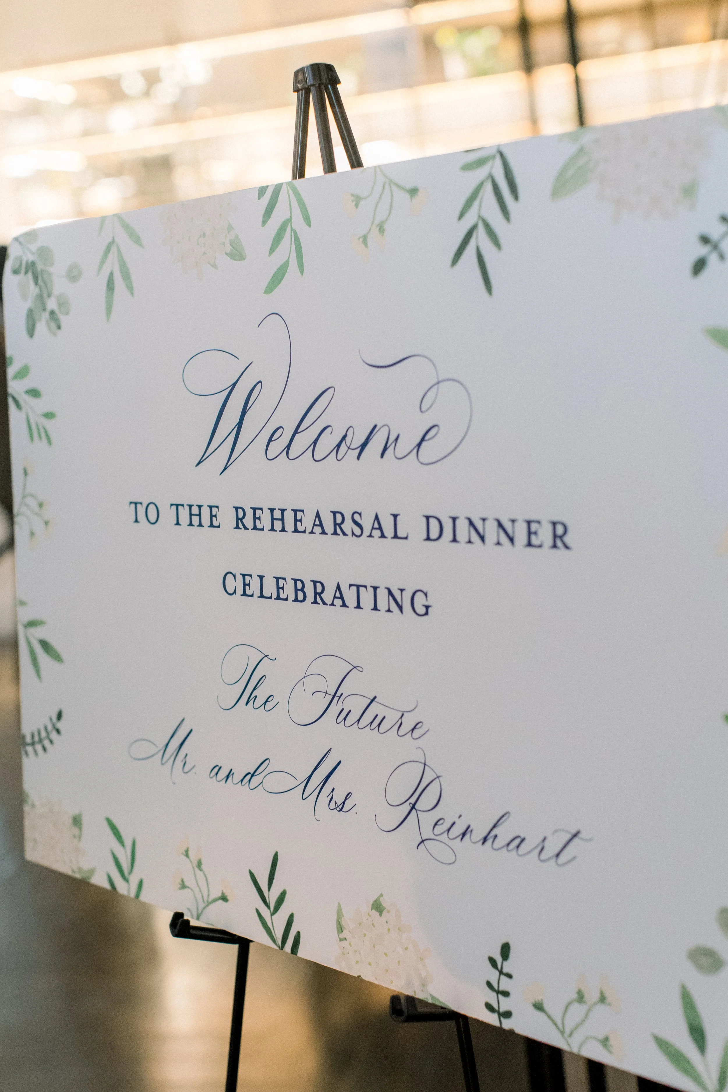 Welcome sign for the rehearsal dinner celebrating Mr. and Mrs. Reinart on an easel, decorated with green leaves and white flowers.