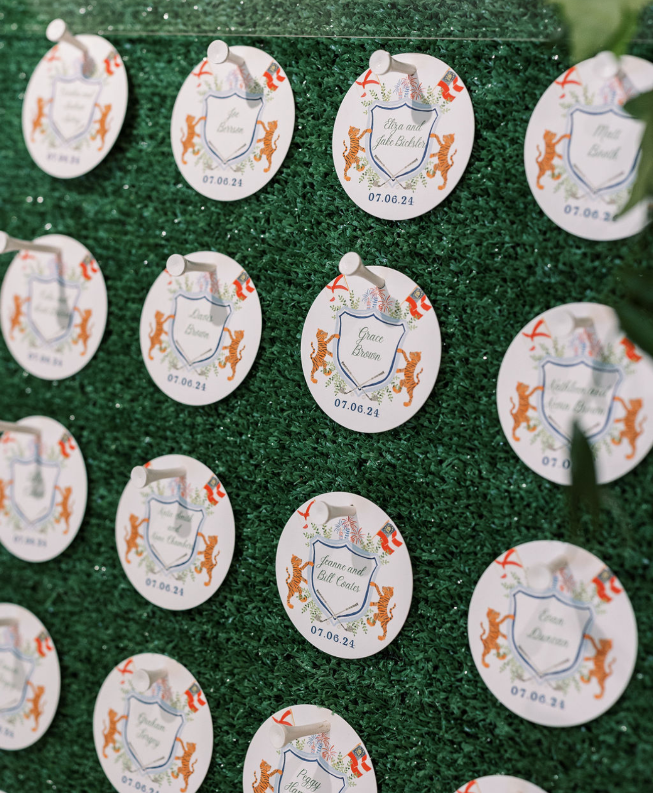 Event seating chart with circular name tags pinned on a green artificial grass backdrop, featuring a crest with orange tigers, red and blue decorative elements, and names of guests with a date of July 6, 2024.
