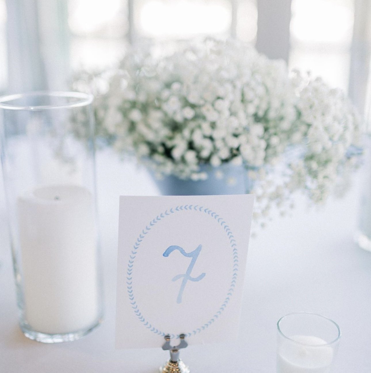 A table setting with a candlestick, a small glass of water, a sign with the letter Z, and a blurred bouquet of white flowers in the background.