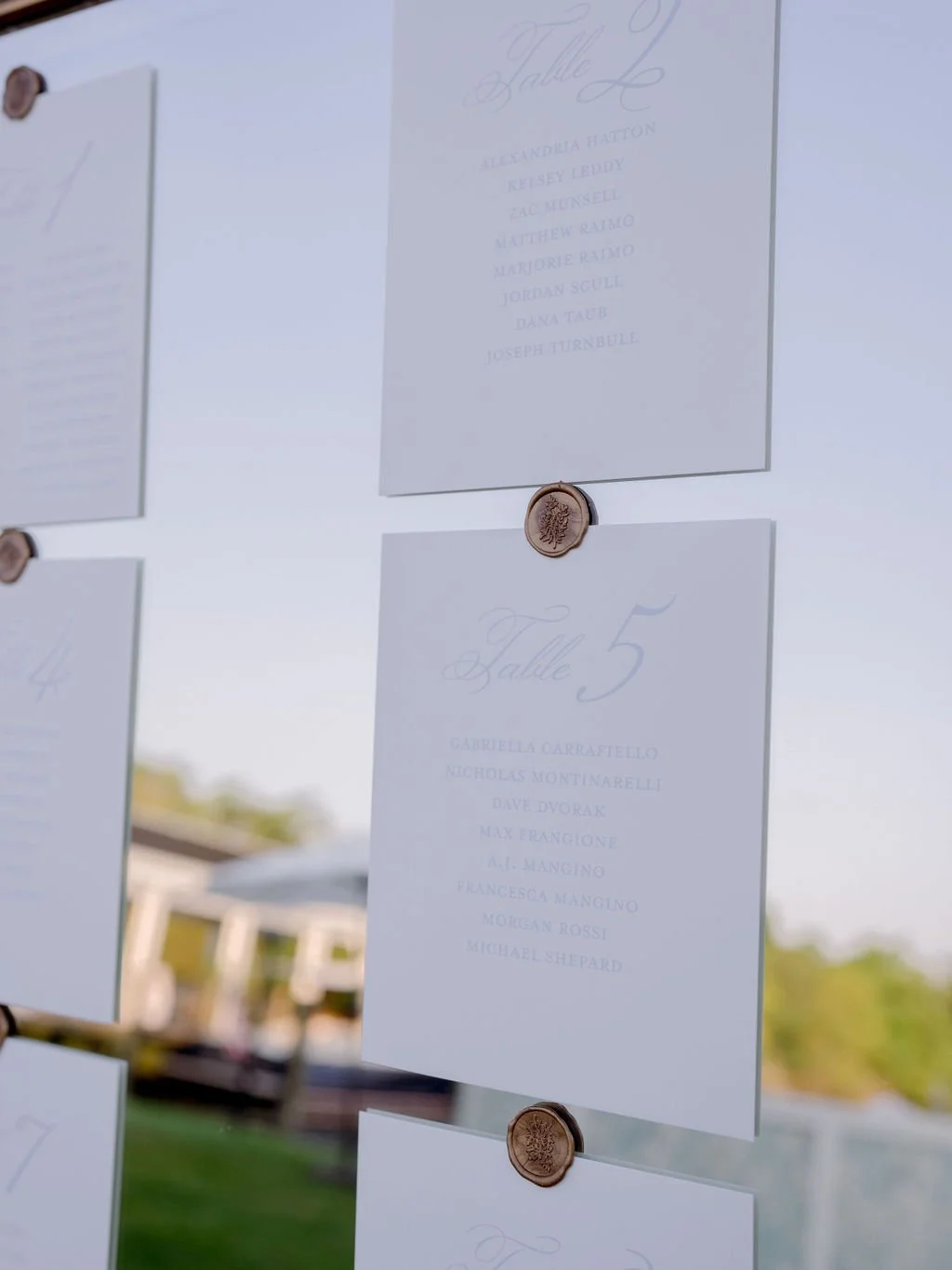 Wedding seating chart with tables labeled in cursive, showing numbered tables and guest names, secured with wax seal stickers, outdoor setting in the background.
