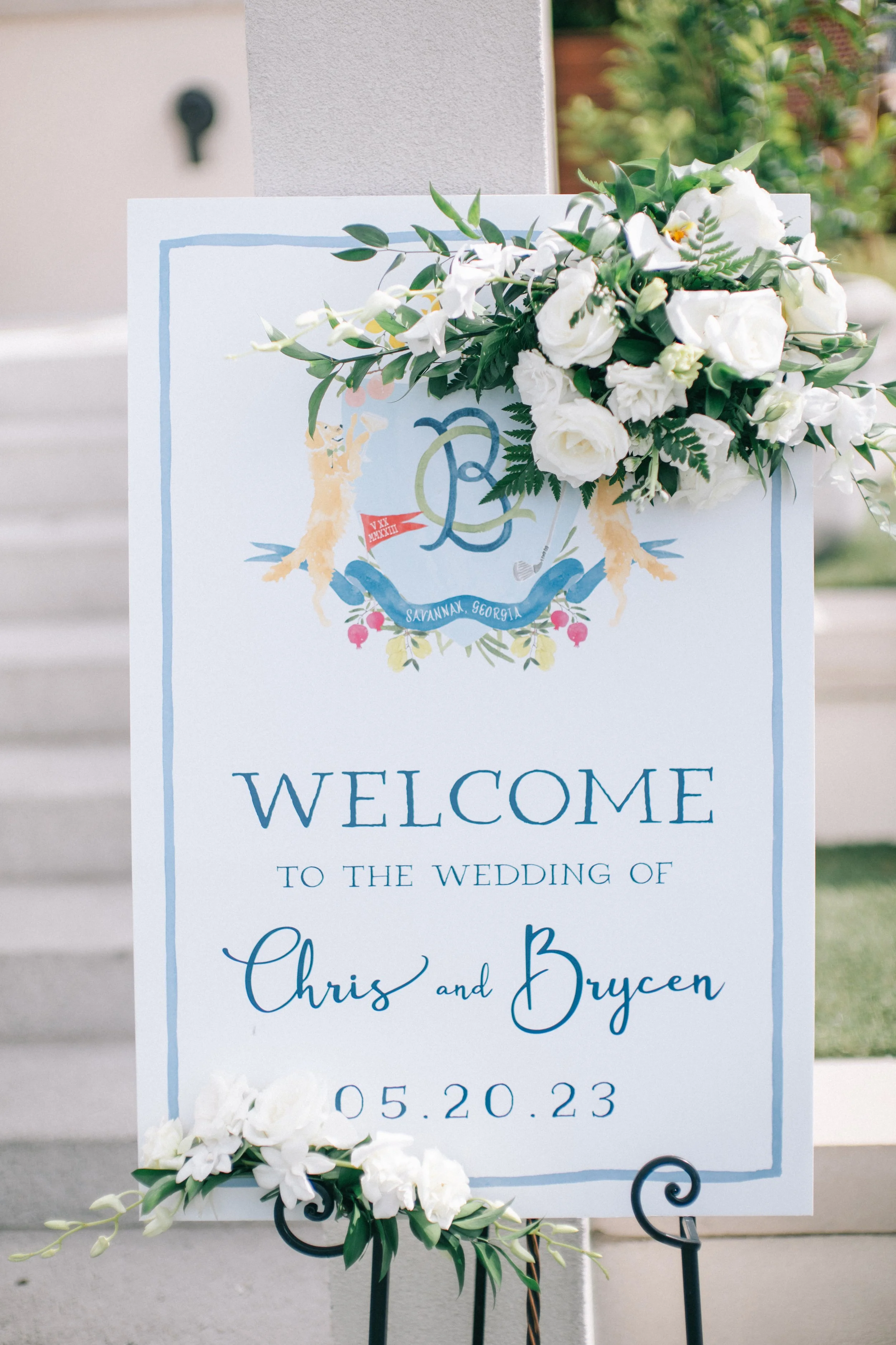 Wedding welcome sign with floral decoration, featuring the names Chris and Bryce and the date May 20, 2023, located outdoors with greenery in the background.