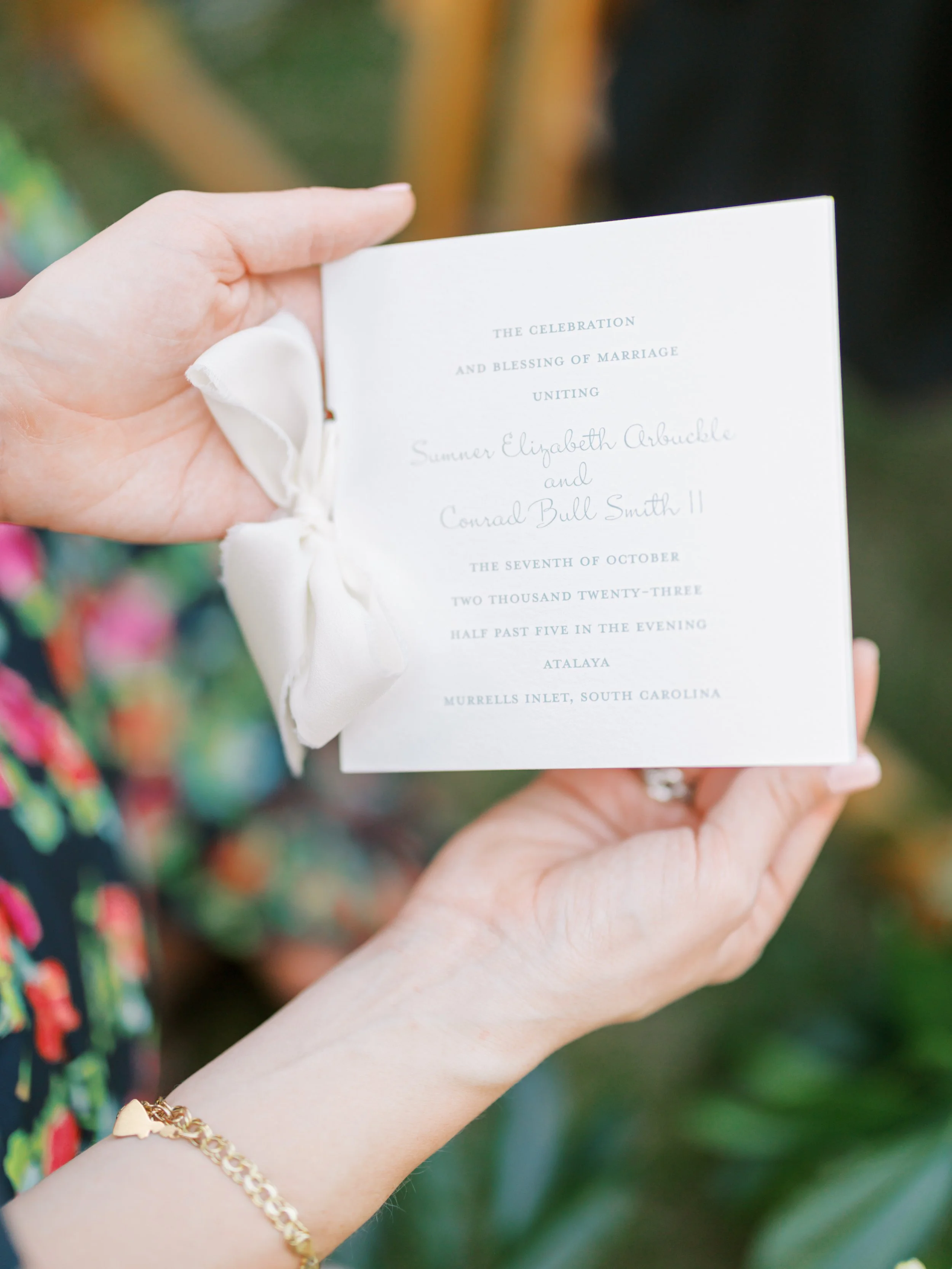 Close-up of a woman holding a wedding program with a white satin bow on the side. The program contains details about a wedding ceremony, including names and date, in light gray and purple text.