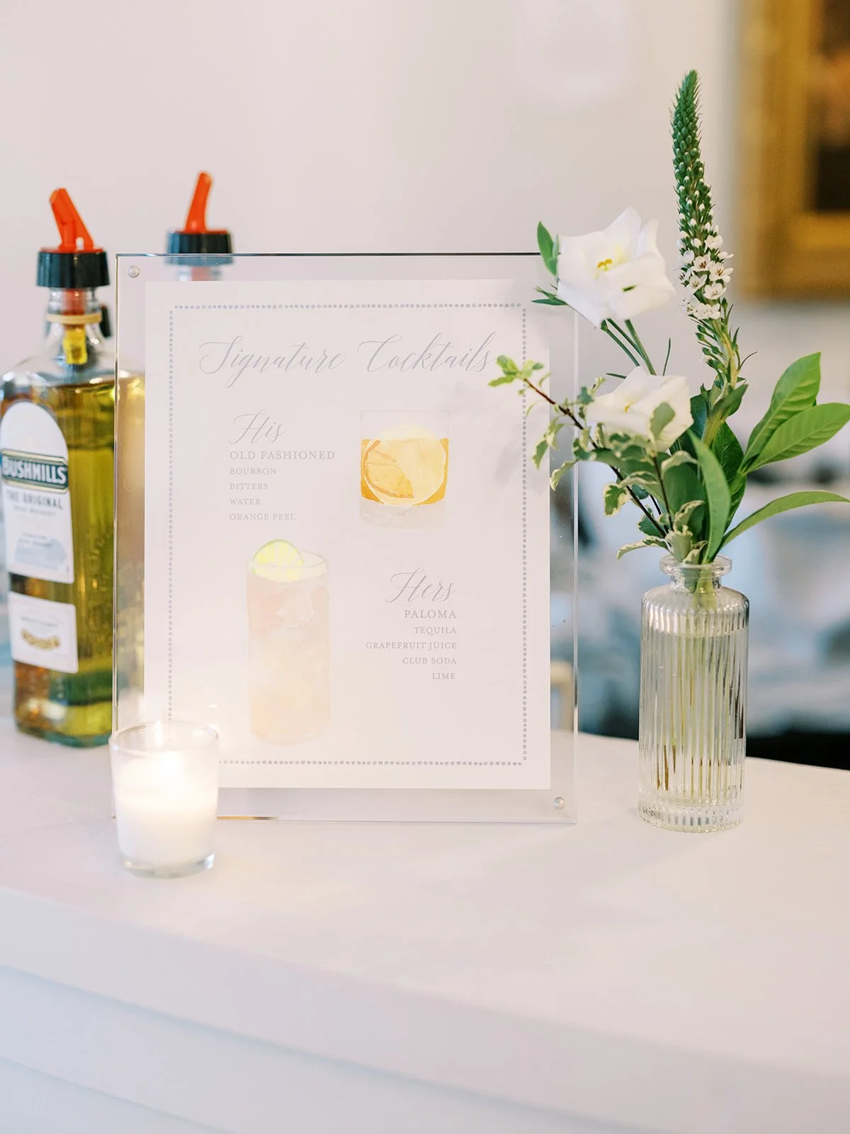 A cocktail menu on a clear stand, with a white candle and a vase with white flowers on a white table.