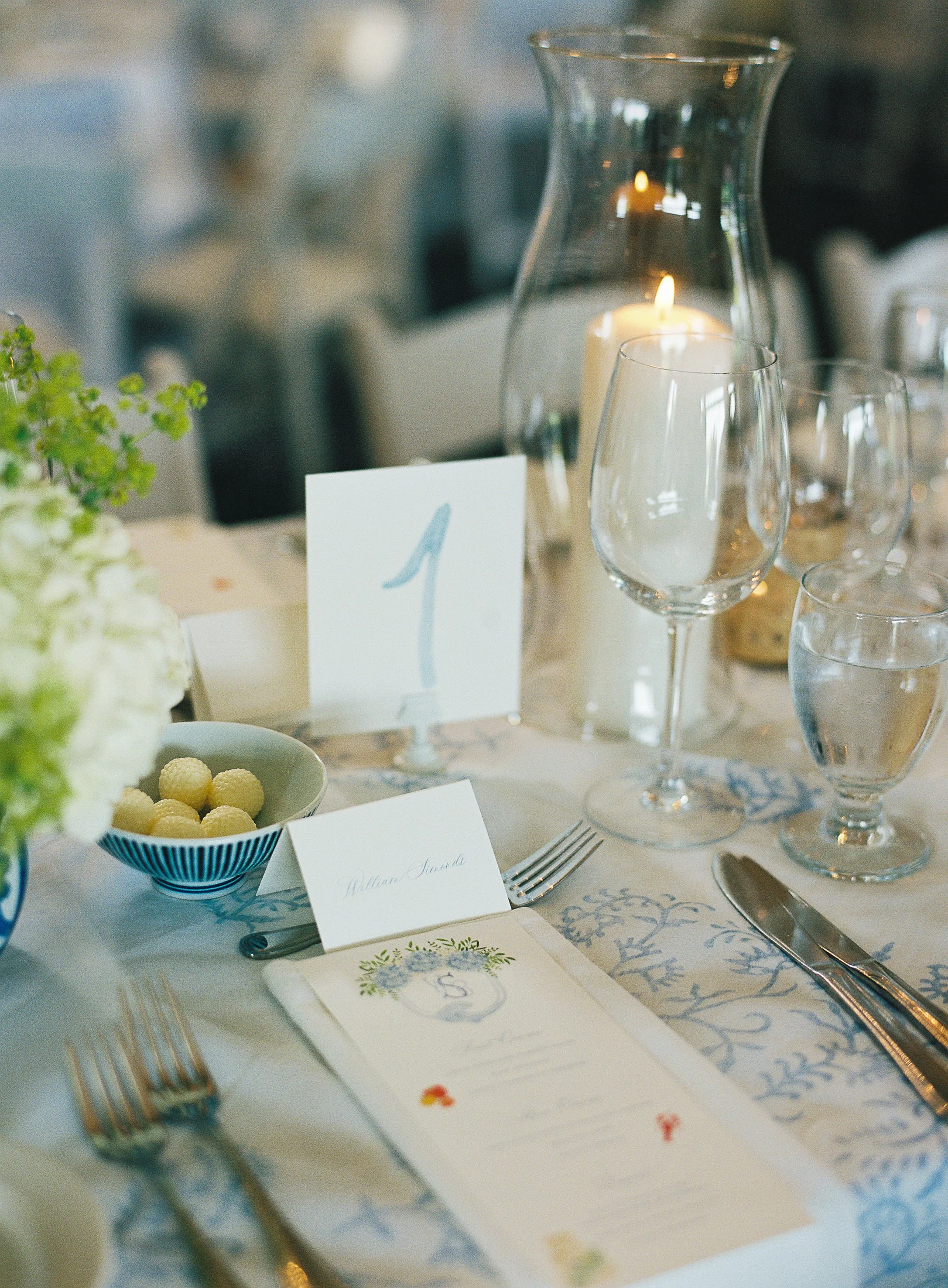 Elegant table setting at a wedding reception with floral decor, water glasses, cutlery, a candle in a glass holder, a table number card, and a menu, on a white tablecloth with blue patterns.