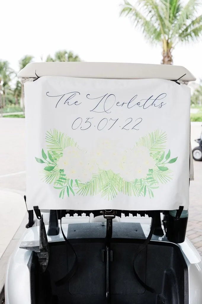 A golf cart with a white cover displaying the event name "The Darlaths" and the date "05.07.22" in elegant script, decorated with green tropical leaves and white flowers.