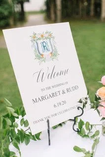 Wedding welcome sign with floral design, featuring initials and the names Margaret and Rudd, dated June 19, 2023, placed on grass likely at an outdoor wedding reception.