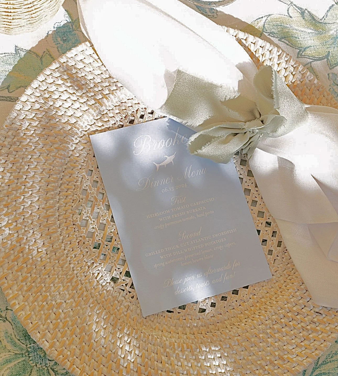 A white dinner menu card with gold lettering, placed on a woven straw hat with a white ribbon, on a table with a floral tablecloth.
