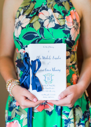 A person wearing a colorful floral dress holding a wedding invitation with a blue ribbon, pearls on their wrist, and the person's arms visible.