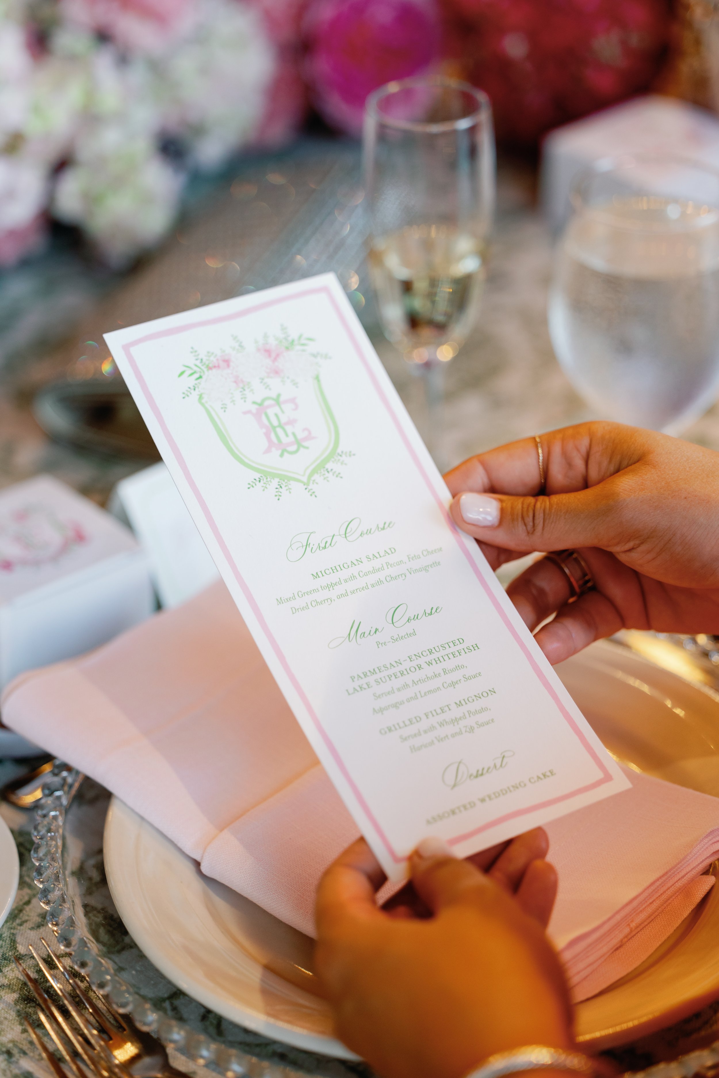 Person holding a wedding menu card at a table with pink napkin, glassware, and floral decorations.