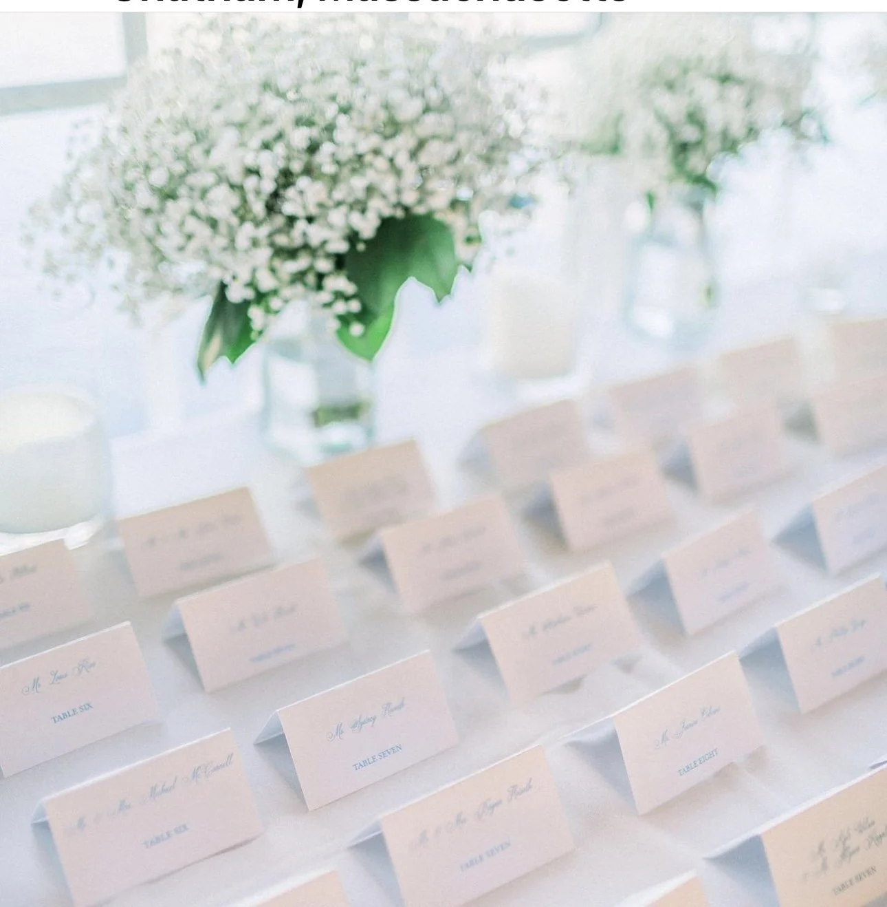 A table with white place cards with blue writing, arranged in rows, with a vase of white flowers and other glass containers in the background.