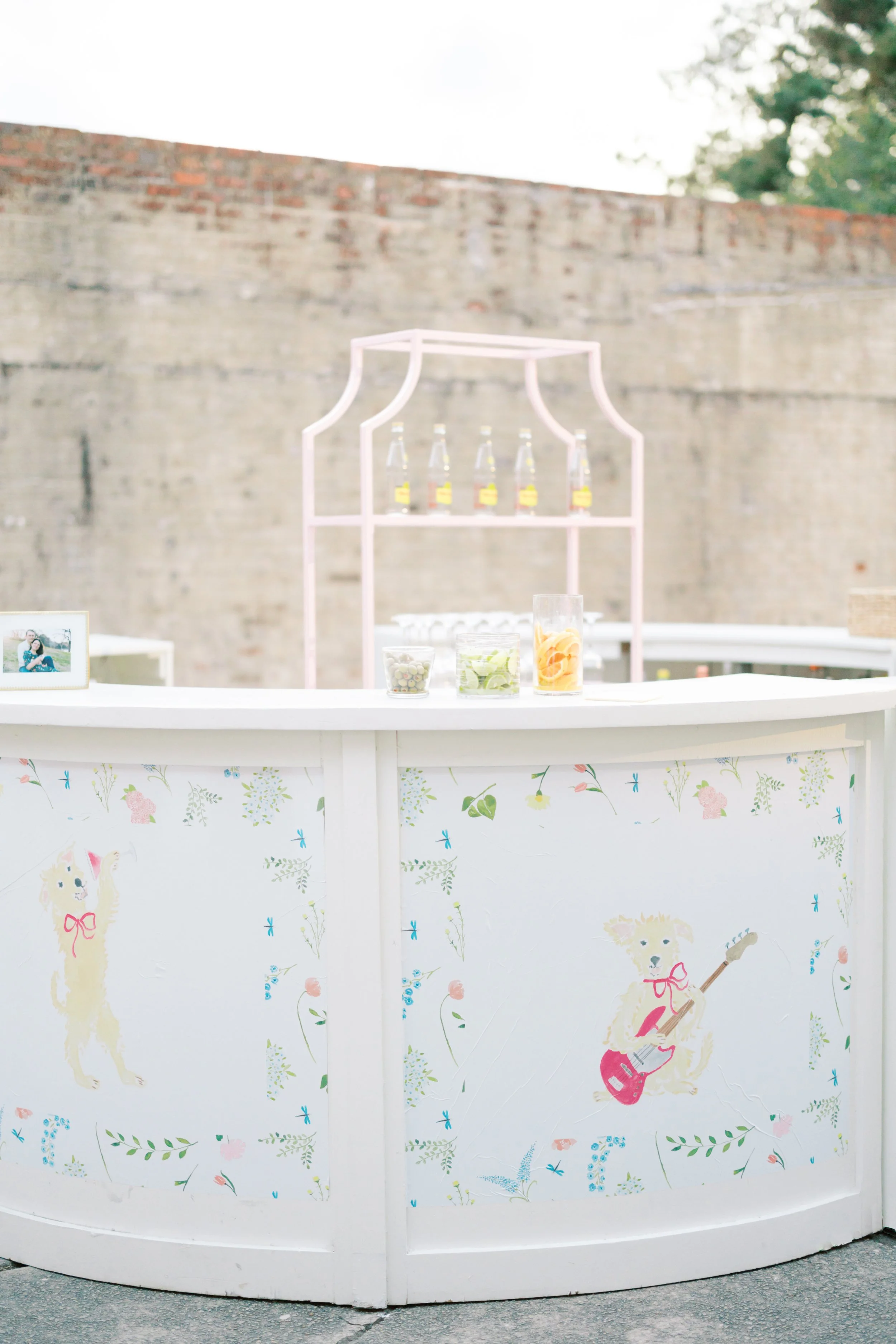 A white bar decorated with cute dog illustrations, including one playing a guitar, outside against a brick wall, with bottles and drinks on top.