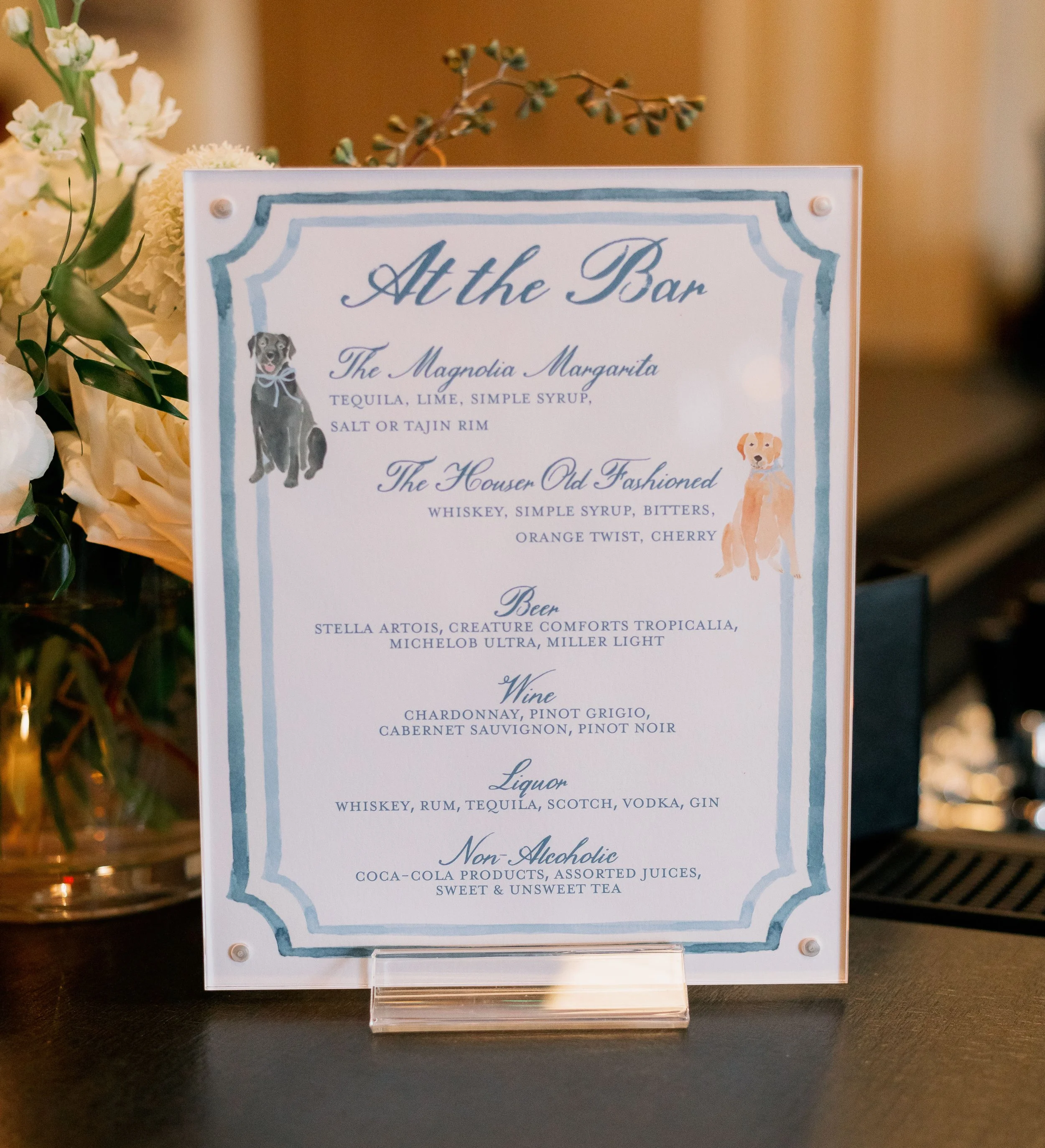 A decorative bar menu sign with illustrations of two dogs, one on each side, displaying drink options including margaritas, old fashioneds, beer, wine, liquor, and non-alcoholic beverages, placed on a table with flowers nearby.