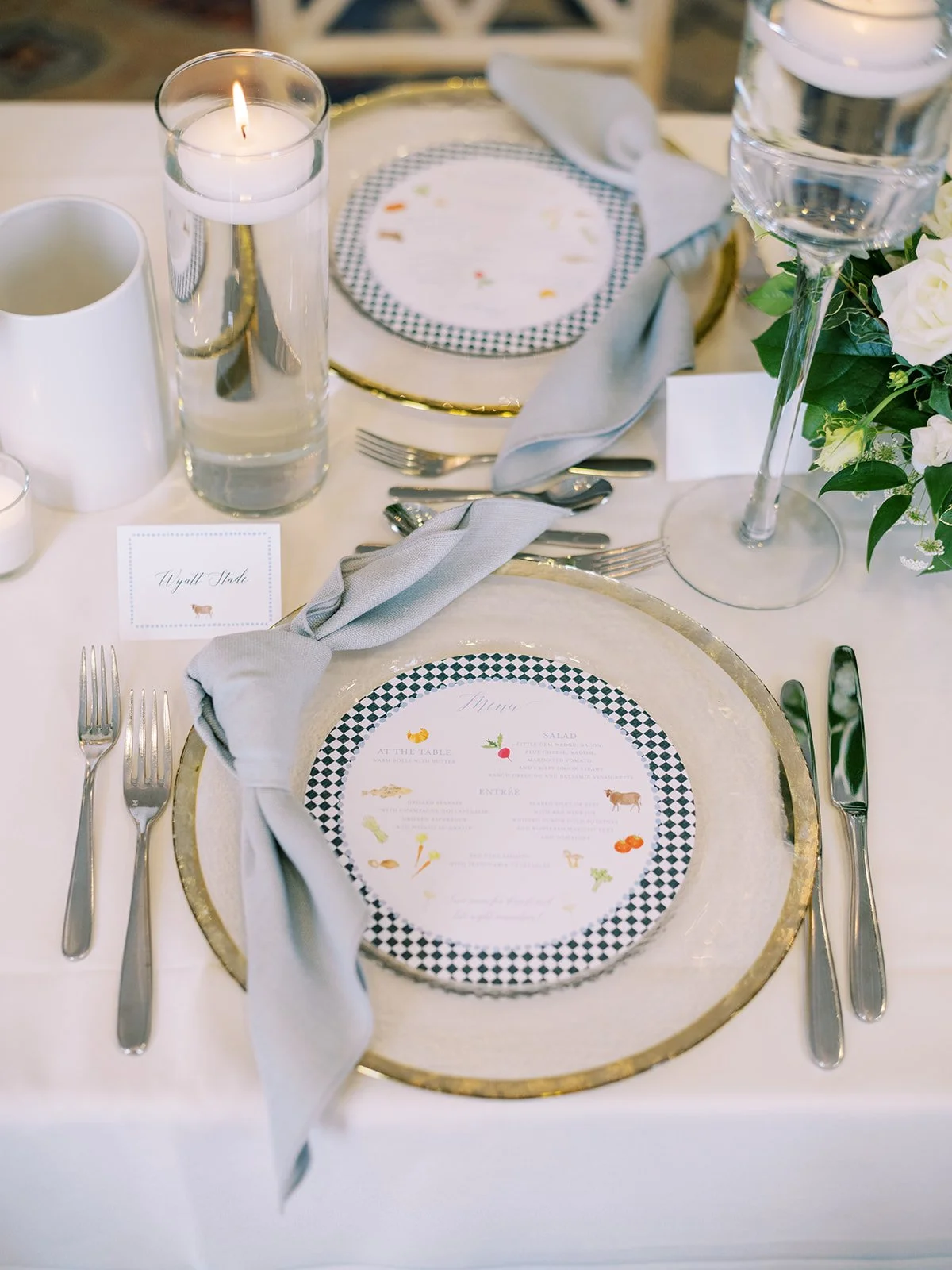 Elegant table setting with a gold-rimmed charger, printed menu, gray napkin, cutlery, water glass with a candle, and white floral centerpiece.