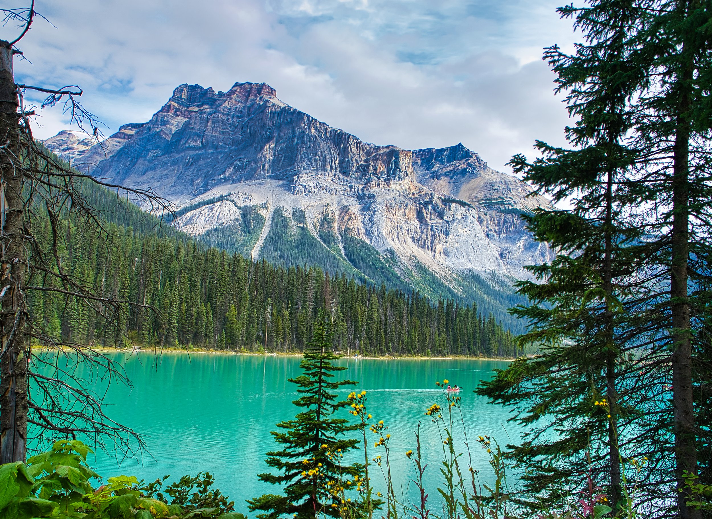Scenic landscape featuring a turquoise lake, surrounded by coniferous trees with a mountain in the background under a partially cloudy sky.