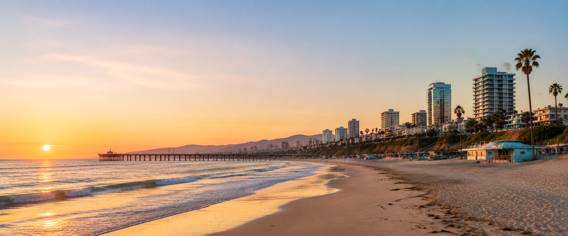 Sunset over the ocean with a pier, sandy beach, high-rise buildings, palm trees, and a few beach houses in the background.