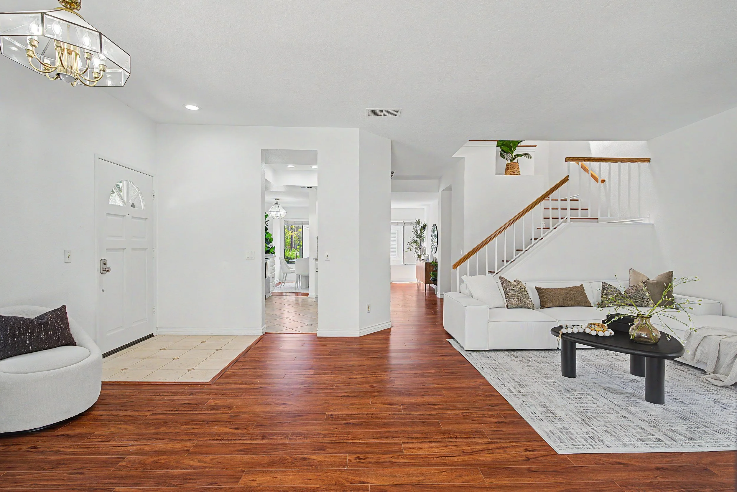 Bright living room with white walls, hardwood floors, white sofa with decorative pillows, round black coffee table with decor, staircase with wooden handrail, entryway with tiled floor, and view into dining area with pendant lighting and a window.