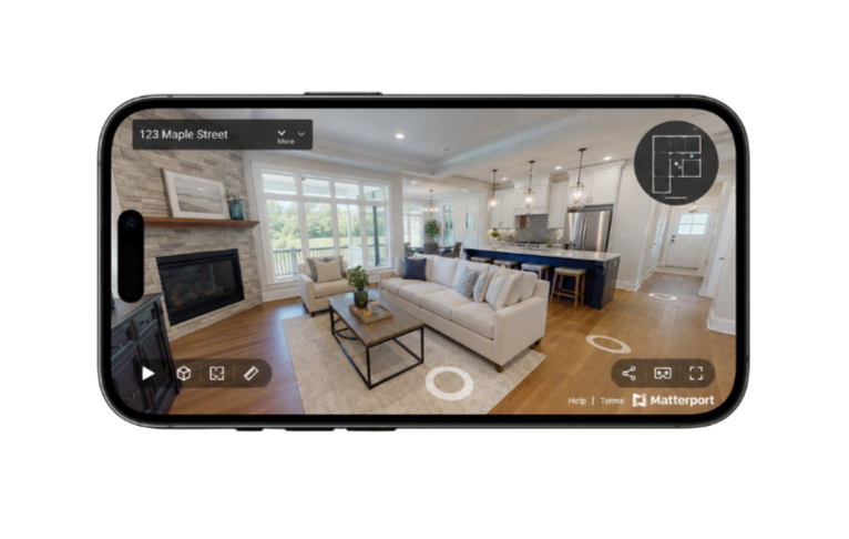 Smartphone displaying a 3D virtual tour of a living room and kitchen inside a modern house, with a fireplace, large windows, and stainless steel appliances.