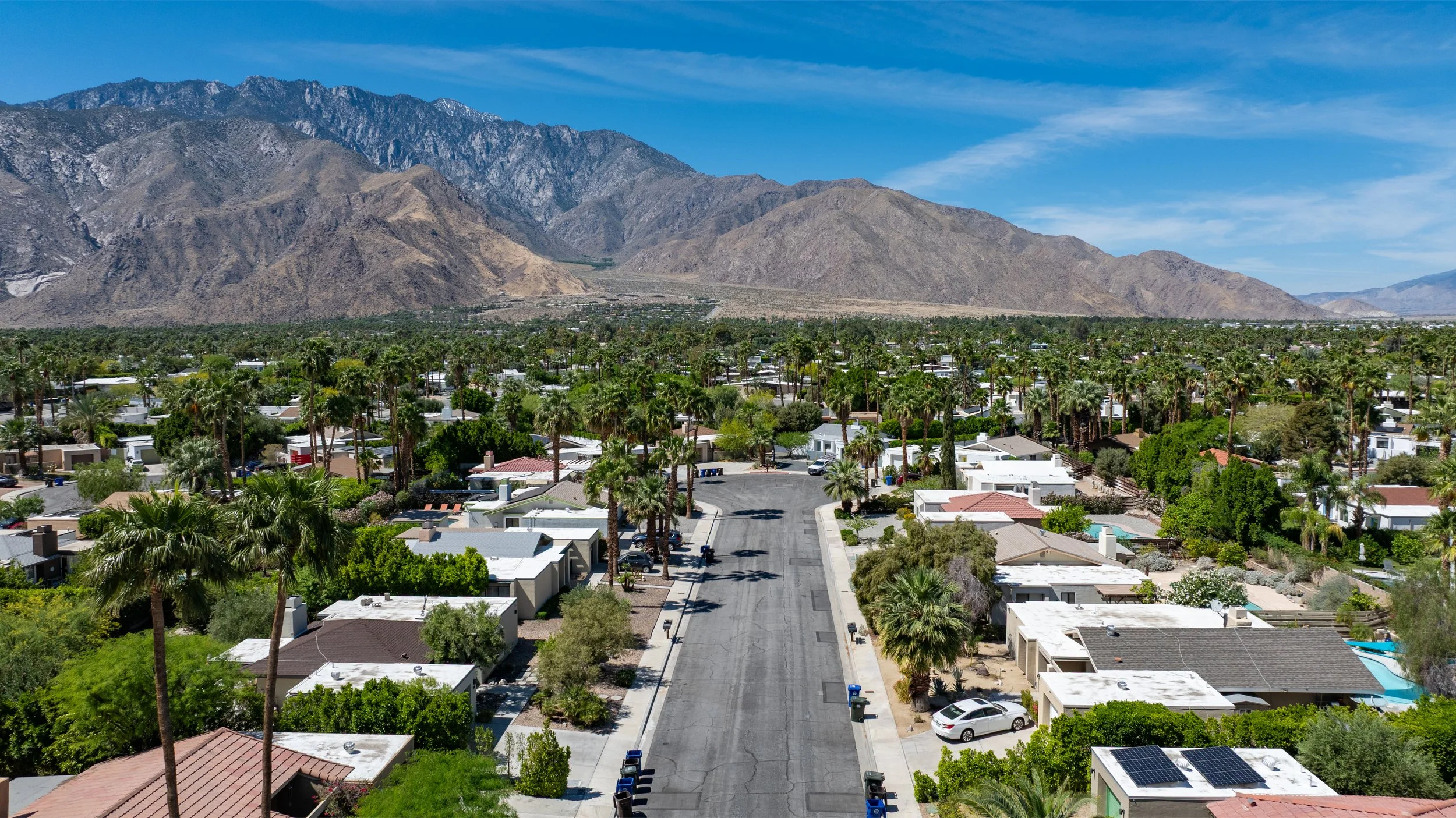 A neighborhood with single-family homes, palm trees, and a mountain range in the background under a cloudy sky.