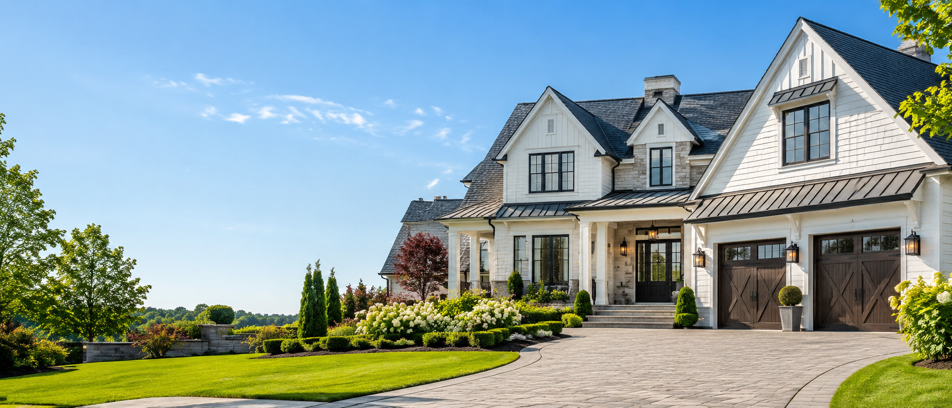 A large, modern white house with black window frames and dark garage doors, surrounded by a well-manicured lawn, colorful shrubs, and trees, under a bright blue sky.