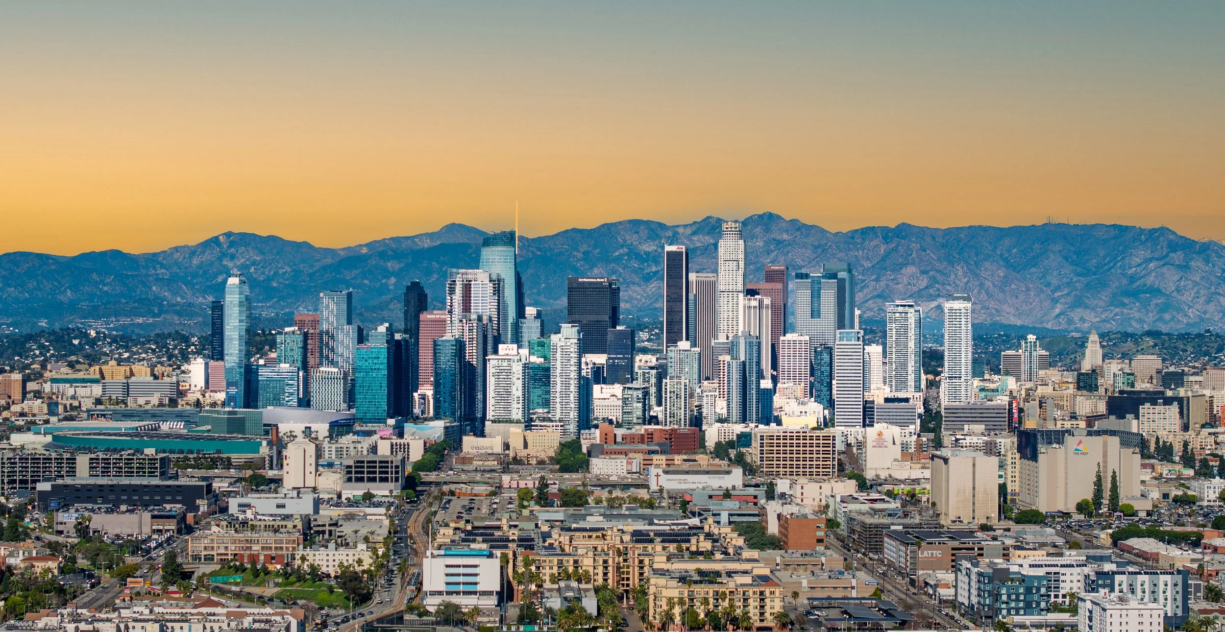 A city skyline featuring numerous tall buildings with mountain ranges in the background during sunset.